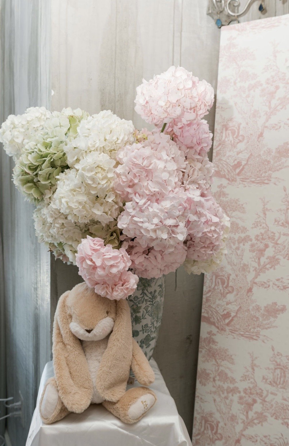 A large bouquet of pink and white hydrangeas in a decorative vase, with a plush bunny stuffed animal sitting in front of it, on a small table covered with a white cloth, with a pink patterned curtain or wall hanging in the background.