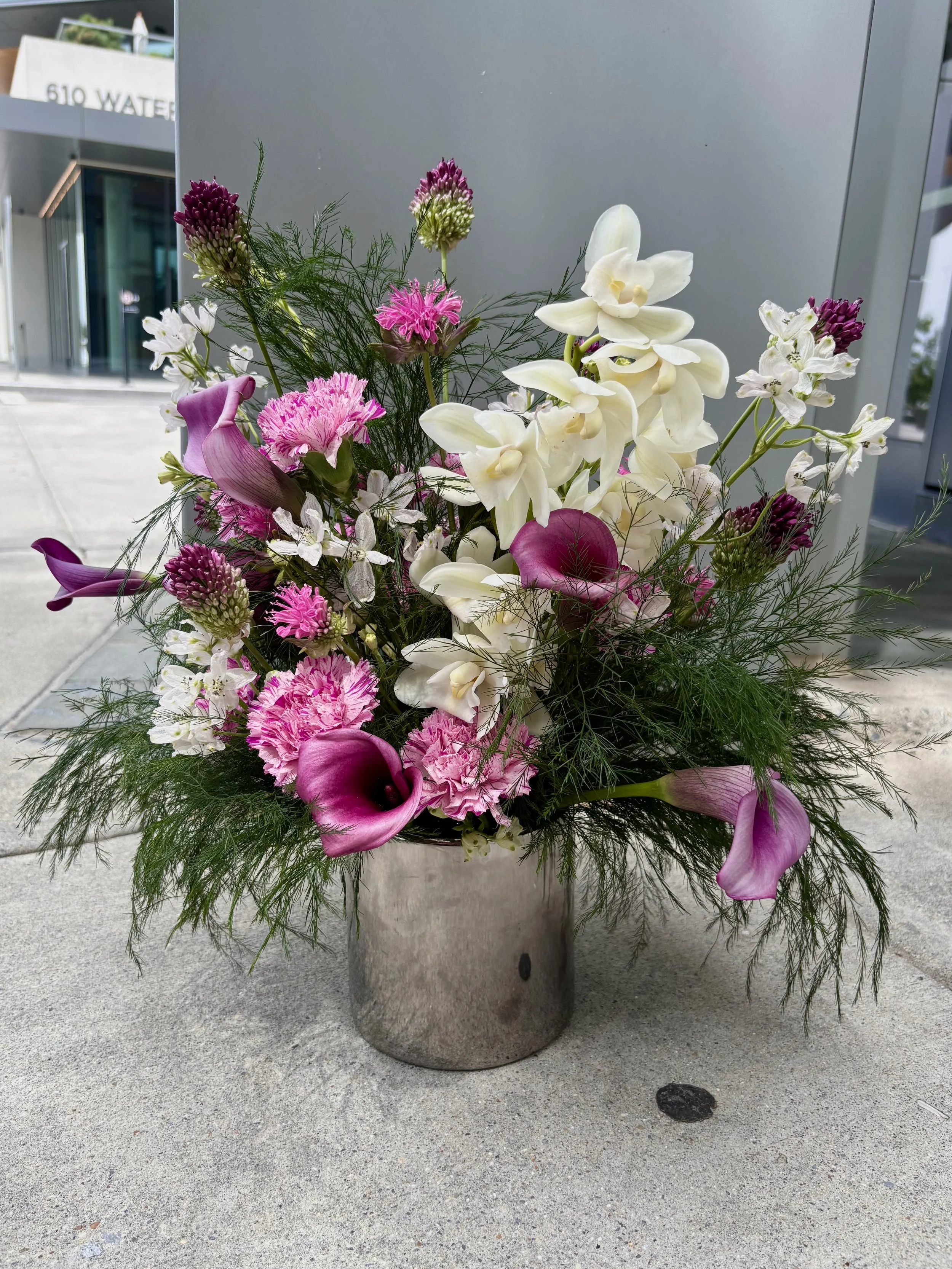 A flower arrangement in a metallic silver vase featuring white orchids, pink carnations, purple calla lilies, and various greenery, placed on a concrete surface outside a building.