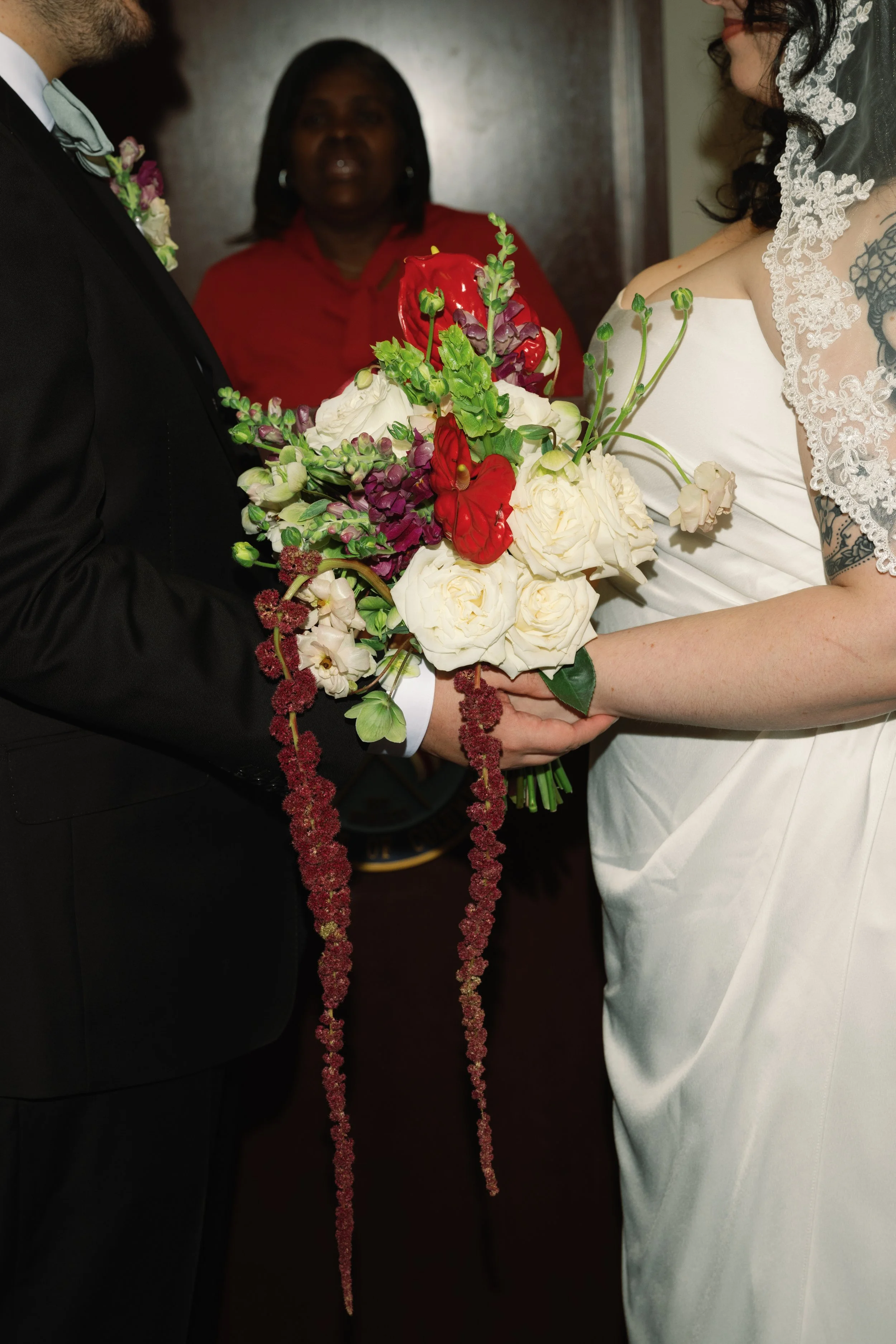 A bride and groom holding a bouquet of white and red flowers during a wedding ceremony, with a woman in a red dress standing in the background.