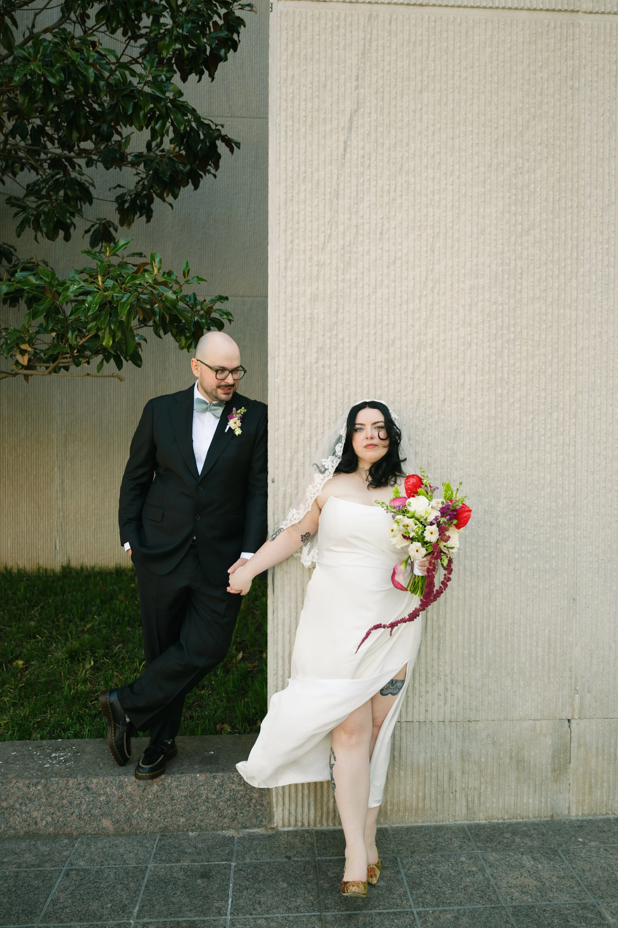 A bride in a white wedding dress and veil holding a bouquet of red, white, and purple flowers, standing next to a groom in a black tuxedo with a bow tie and boutonniere. The groom is leaning against a large concrete wall, holding hands with the bride, who is showing her tattooed leg through a high slit in her dress. They are outdoors with green grass and a tree in the background.