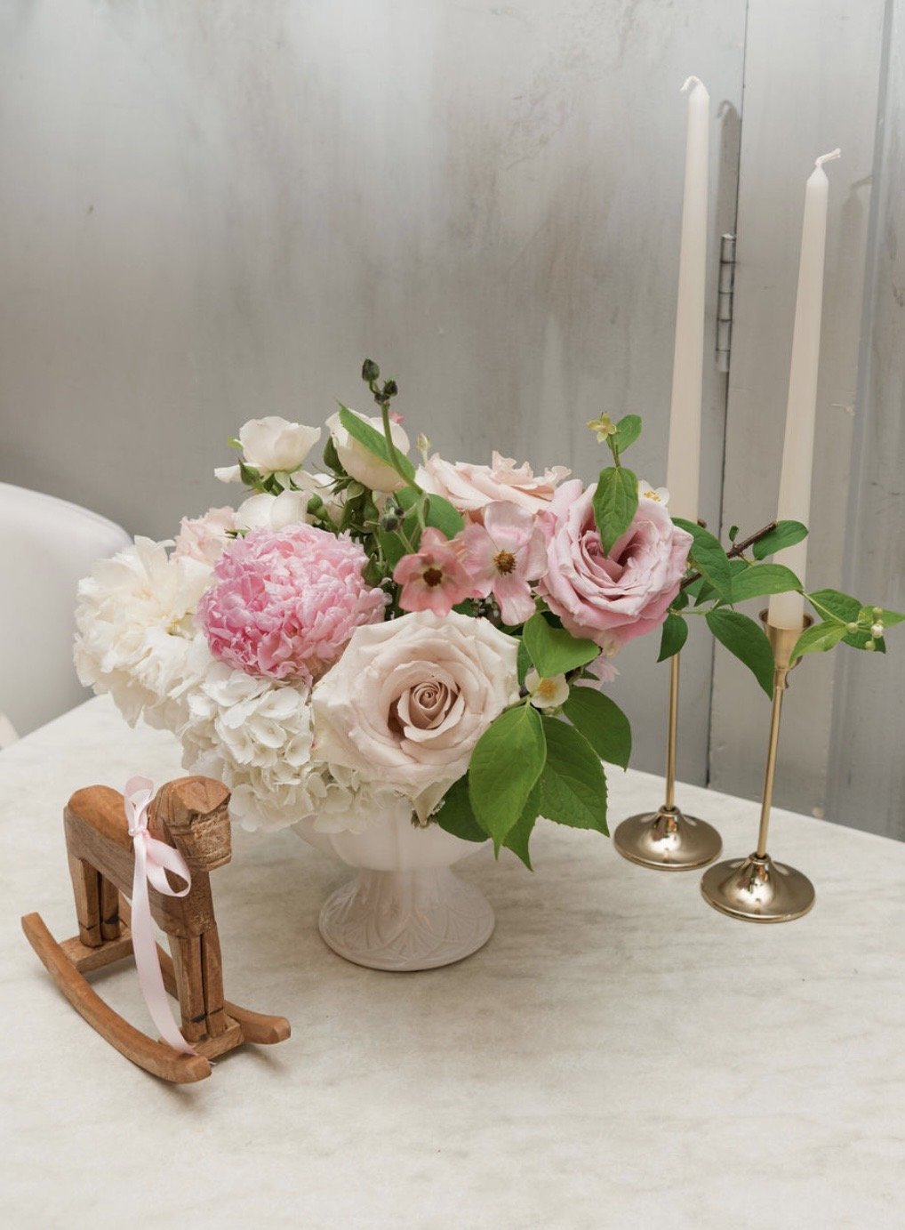 A flower arrangement with pink and white roses, peonies, and greenery in a white vase, placed on a table with two gold candlesticks holding white candles and a small wooden sled with a pink ribbon.