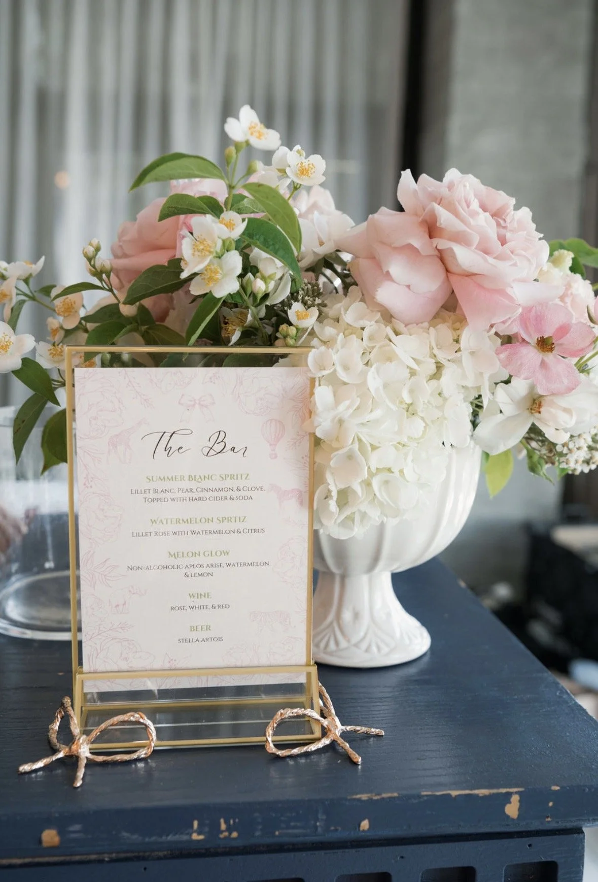 A large white vase filled with pink and white flowers, including roses and hydrangeas, placed on a dark table. In front of the flowers, there is a stylish menu sign with the heading 'The Bar' listing various drinks, including spritzes, watermelon glow, wine, and beer.