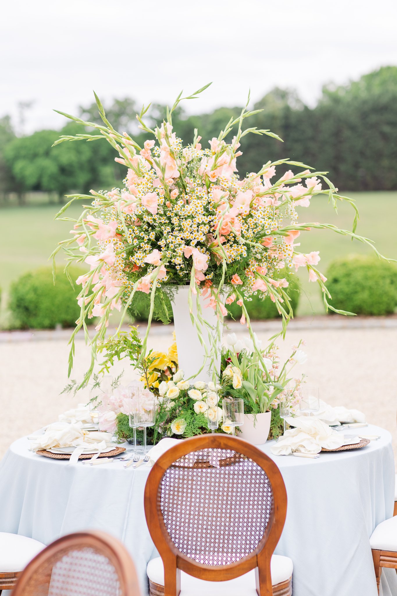A round table decorated with an elaborate floral centerpiece of pink and white flowers and greenery, set outdoors with a white tablecloth, plates, napkins, glassware, and chairs with wooden frames and woven cane backs. The background shows a grassy area and trees under a cloudy sky.