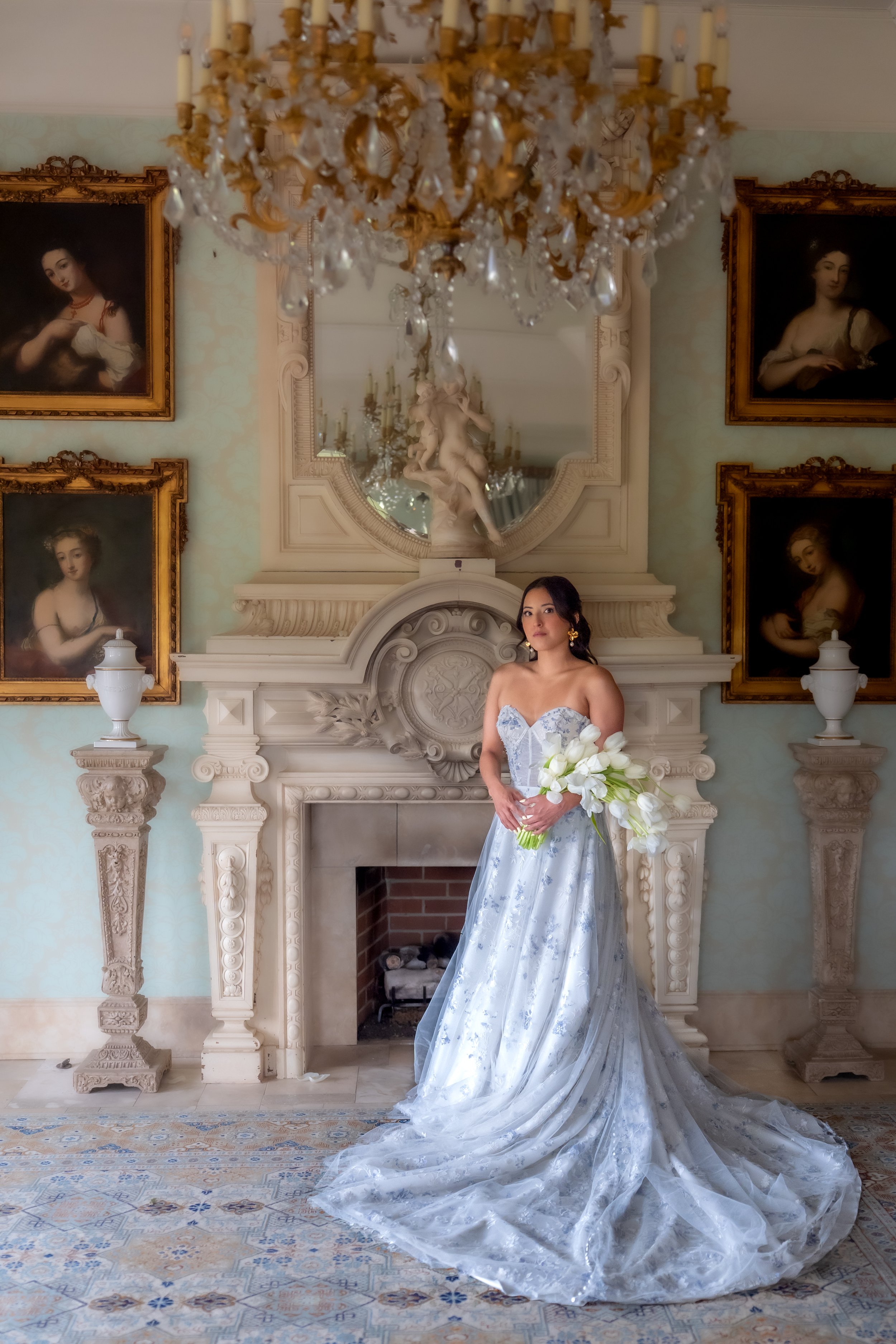 A woman in a strapless white and blue floral wedding gown holding a bouquet of white tulips, standing in front of an ornate fireplace with a large mirror and a chandelier in a classic, elegant room decorated with framed paintings.