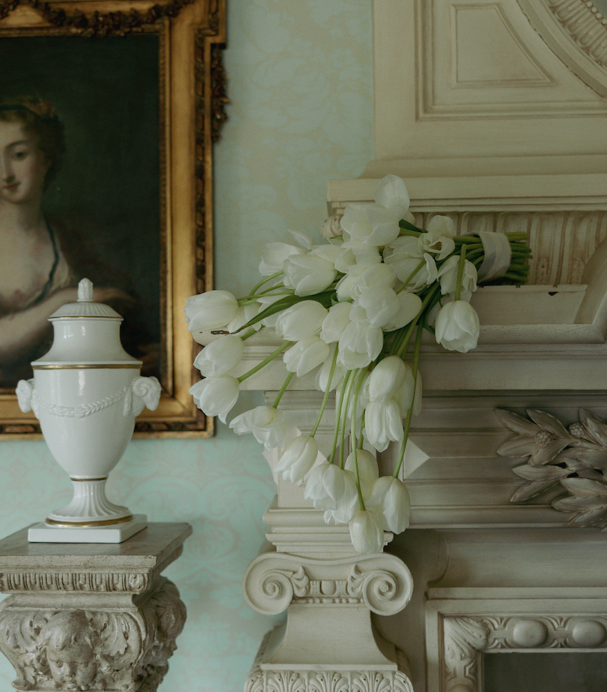 White tulips placed on a decorative fireplace mantel in a vintage styled room with ornate furniture and a portrait painting in the background.