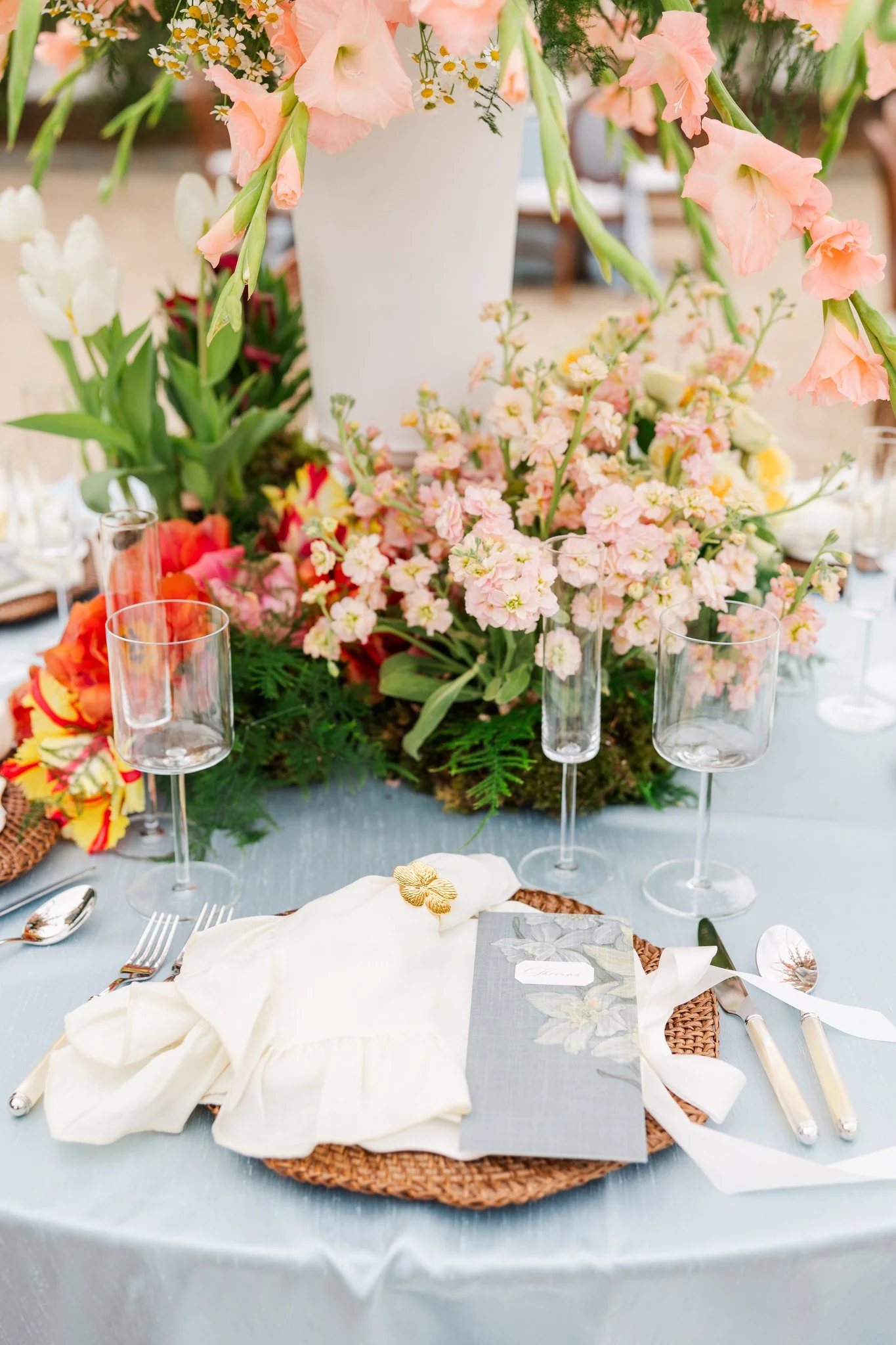 A table setting with a floral centerpiece featuring pink and white flowers, empty wine glasses, and place settings with cream napkins, silverware, and a menu or card on a woven placemat.