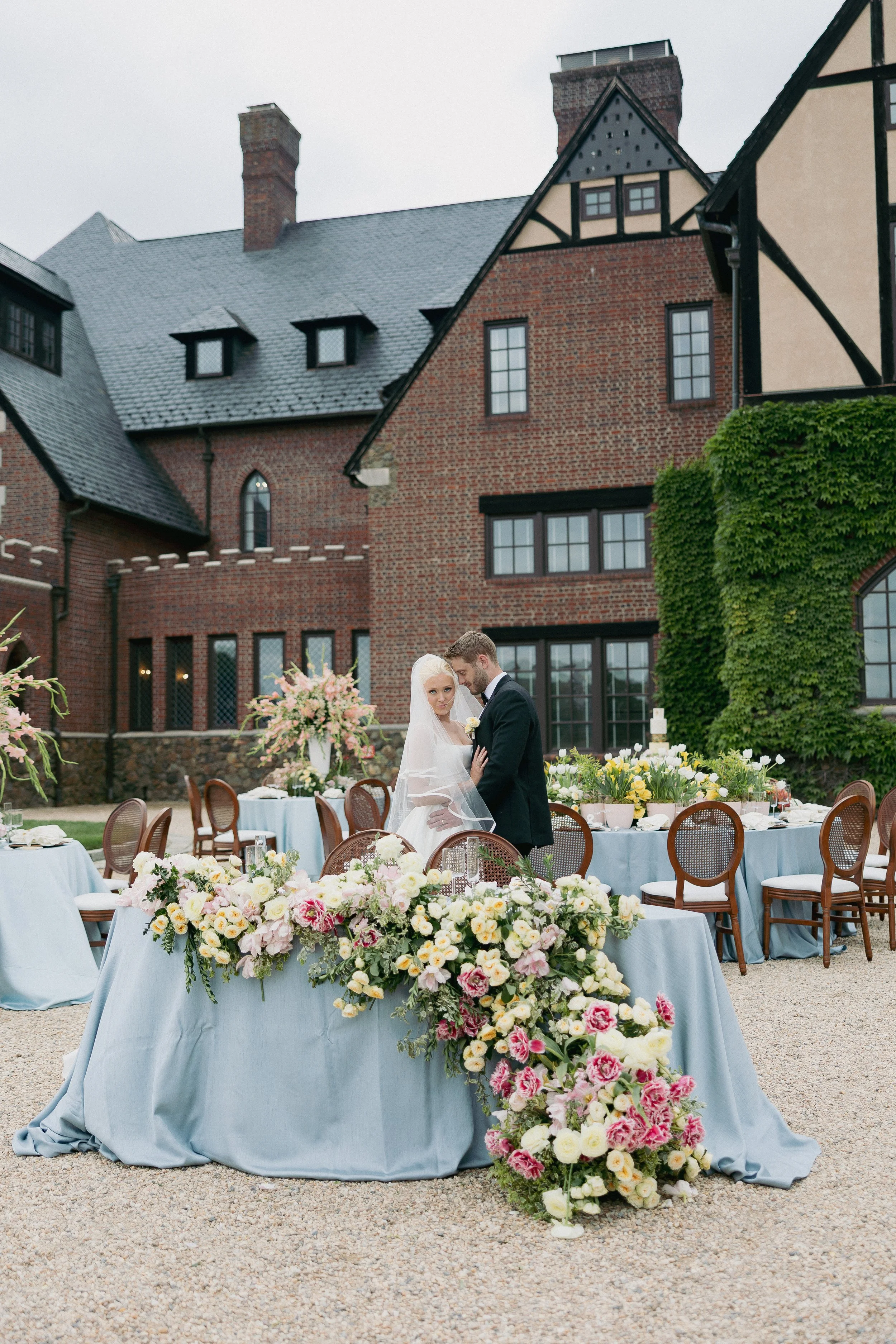 Wedding reception outdoors with a bride and groom embracing, decorated tables with pastel-colored flowers, set against a historic brick building with large windows and ivy growth.