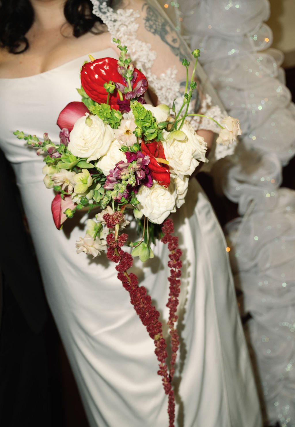 A bride in a white wedding dress holding a cascading bouquet of white roses, red anthuriums, pink flowers, and greenery, with a blurred background featuring a woman in a silver dress.