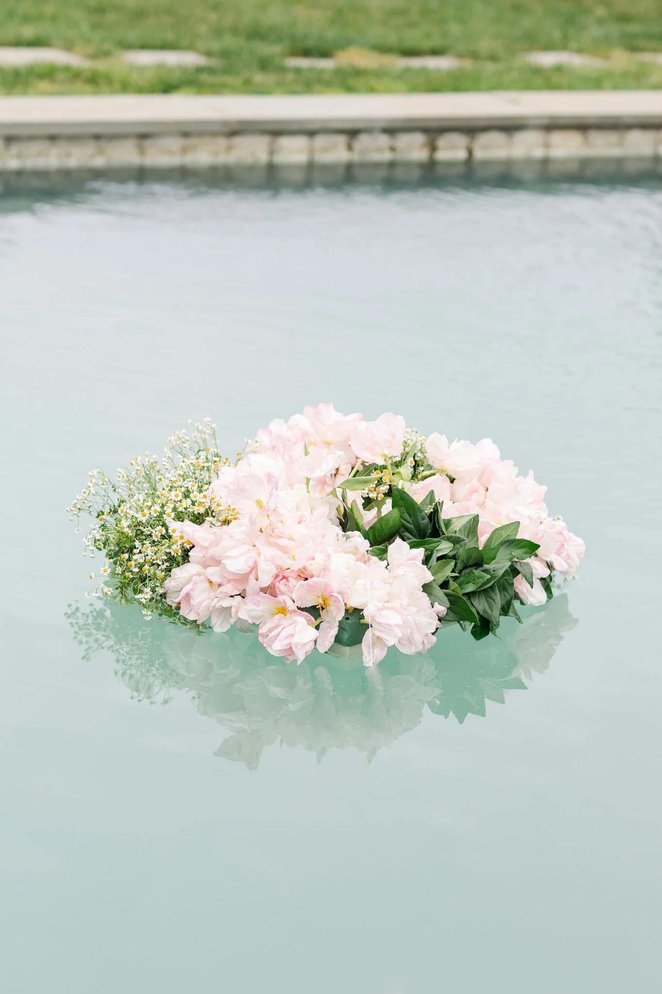 Pink and white hydrangea bouquet with small white daisies and greenery reflected on a glass surface with a blurred grassy outdoor background.