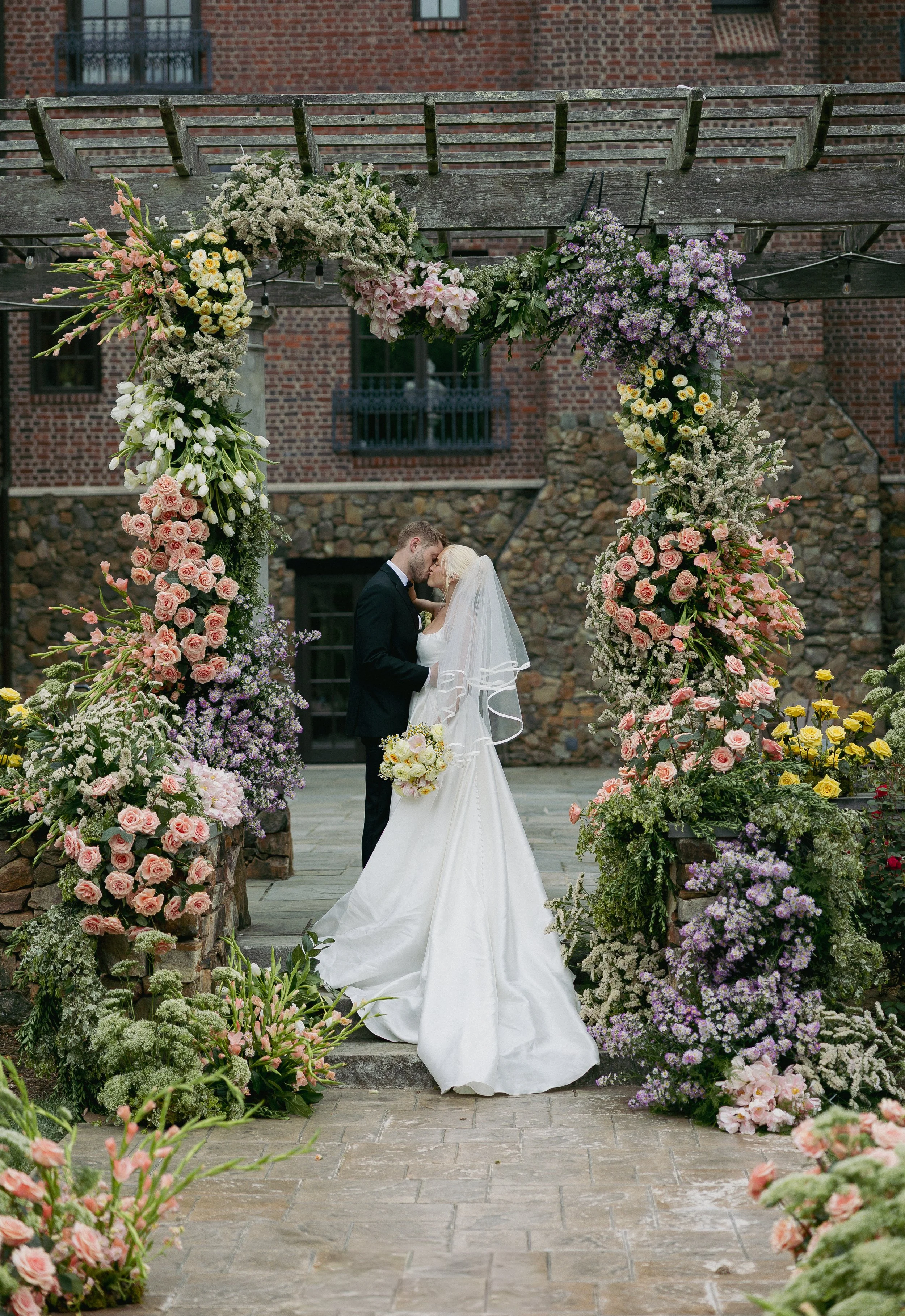 A bride and groom sharing a kiss under a decorated floral wedding arch outdoors, with brick building in the background.