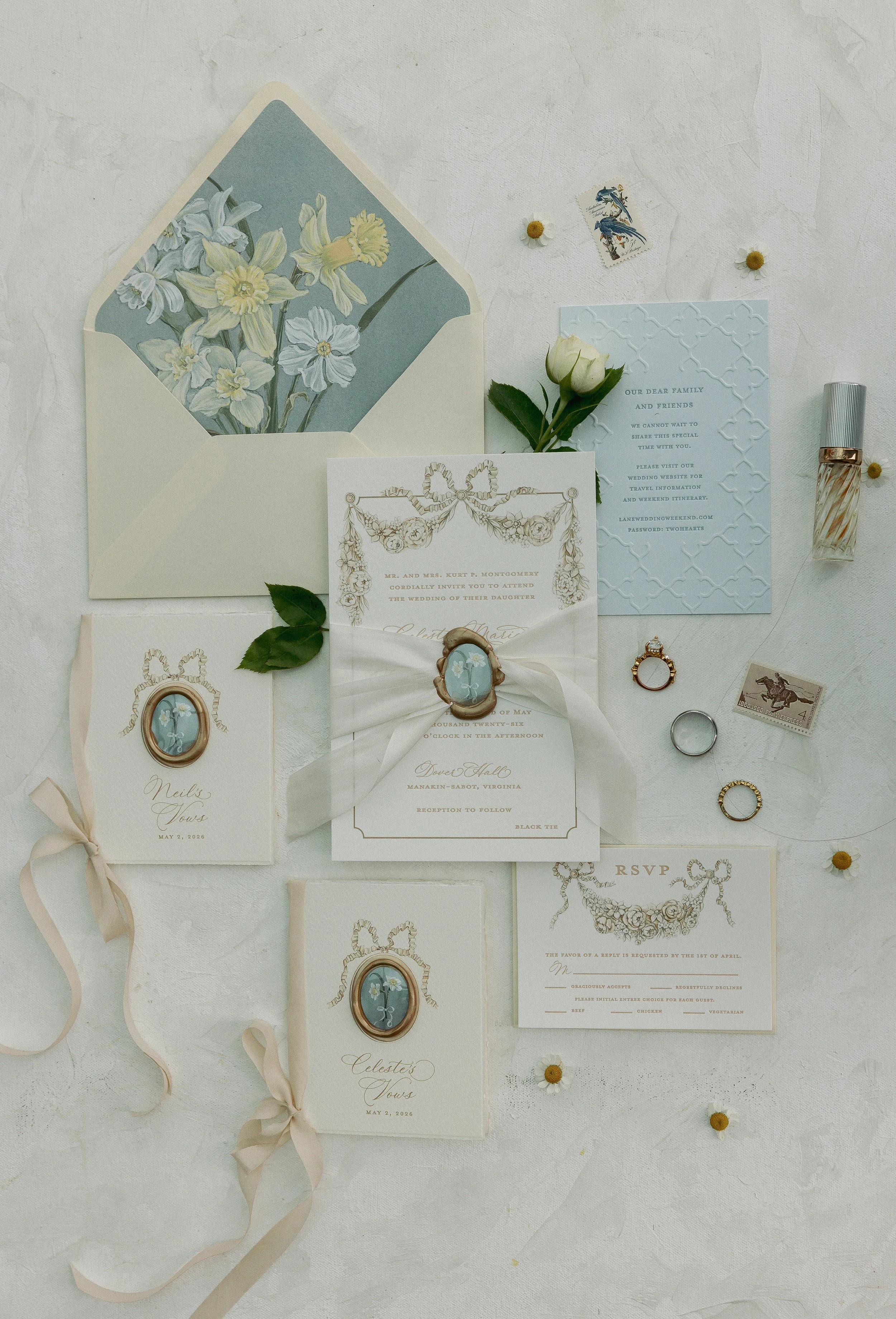 Set of wedding invitations and accessories including a white floral invitation, a blue patterned invitation, a white invitation with gold framing, a small card with a floral design, a tiny envelope, a ring, a necklace, a perfume bottle, a white rose, small yellow flowers scattered around, and an oval cameo frame with a blue gemstone.