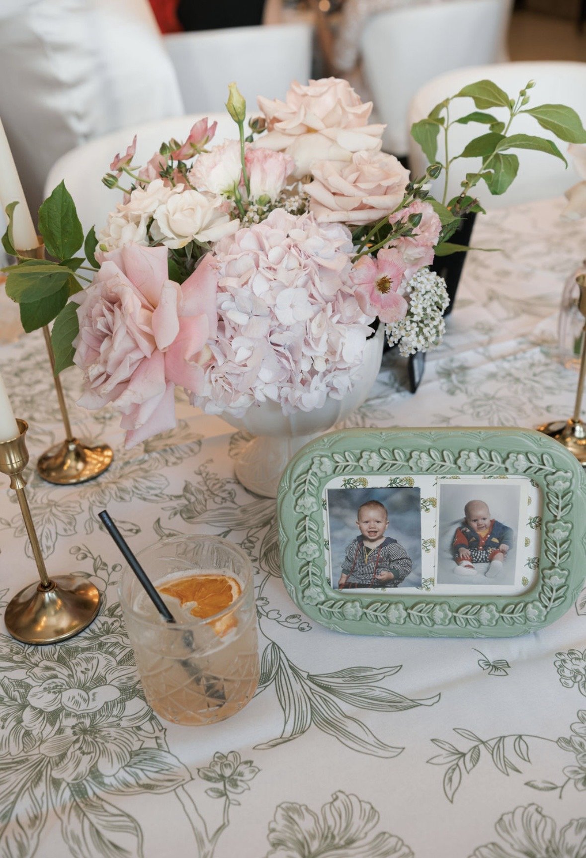 A floral centerpiece with pink and white flowers on a decorated table, next to a framed photo of two young children, and a drink with an orange slice and a straw in the foreground.