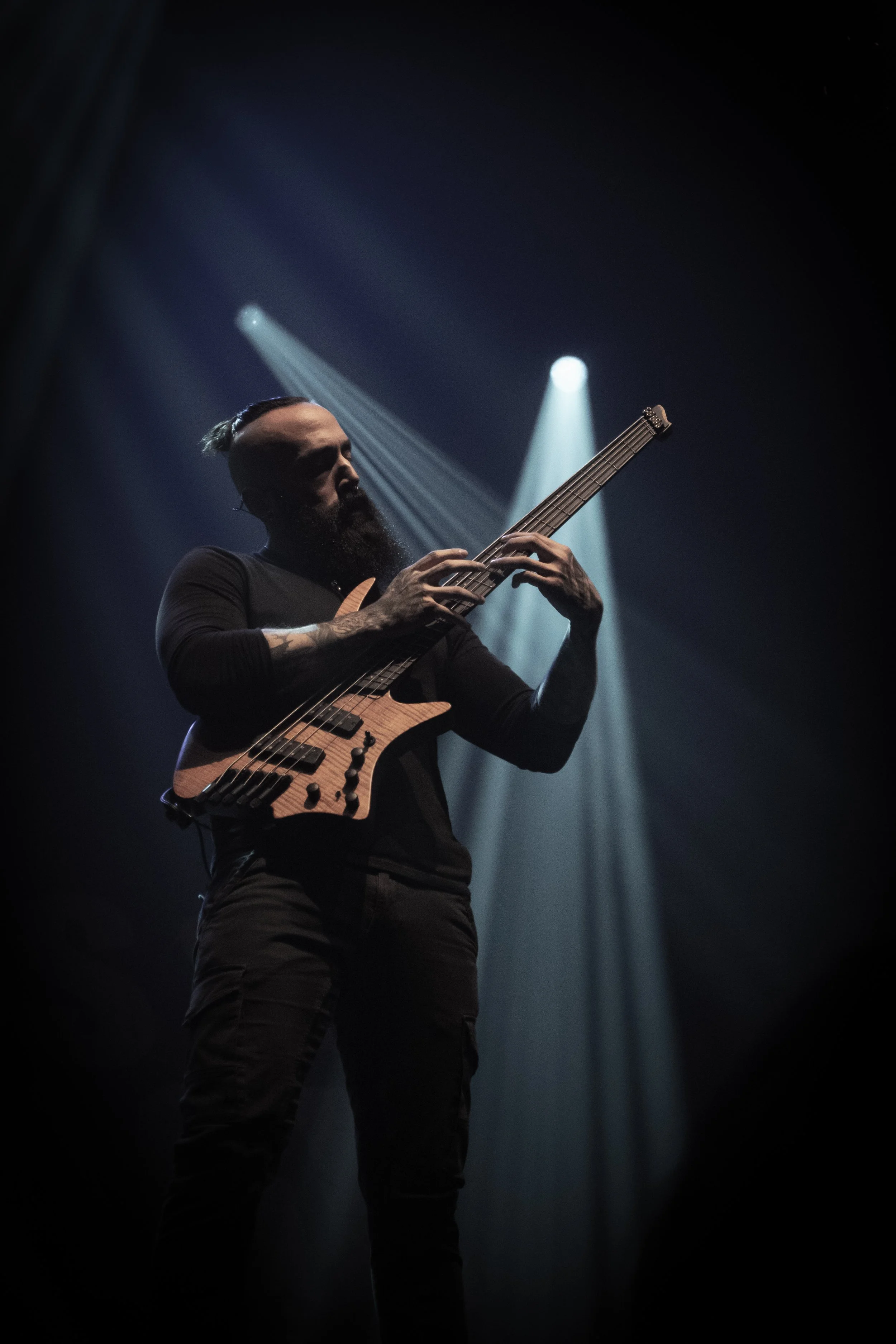 A musician with a beard and tattoos plays an electric guitar on stage illuminated by dramatic lighting.