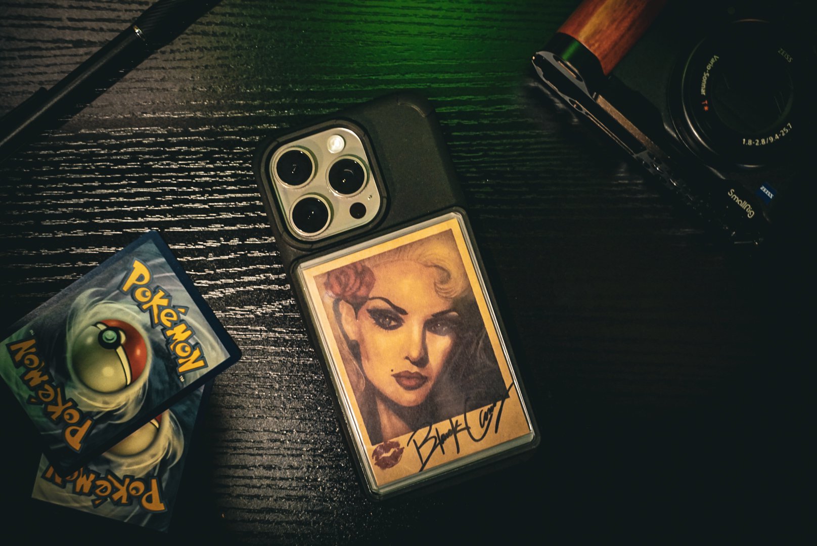 A black table with a portrait of a woman signed 'BlondeCut' on a phone case, a vintage camera, a spring loaded Imposter card, and a pen.