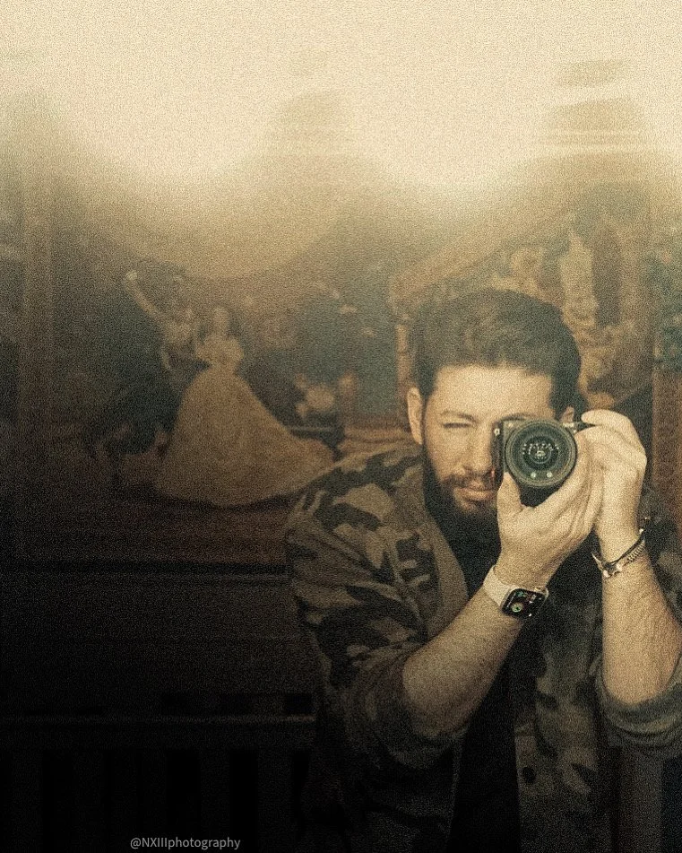 Self-portrait of a man taking a photo with a digital camera in front of a mirror, with a classical painting in the background.