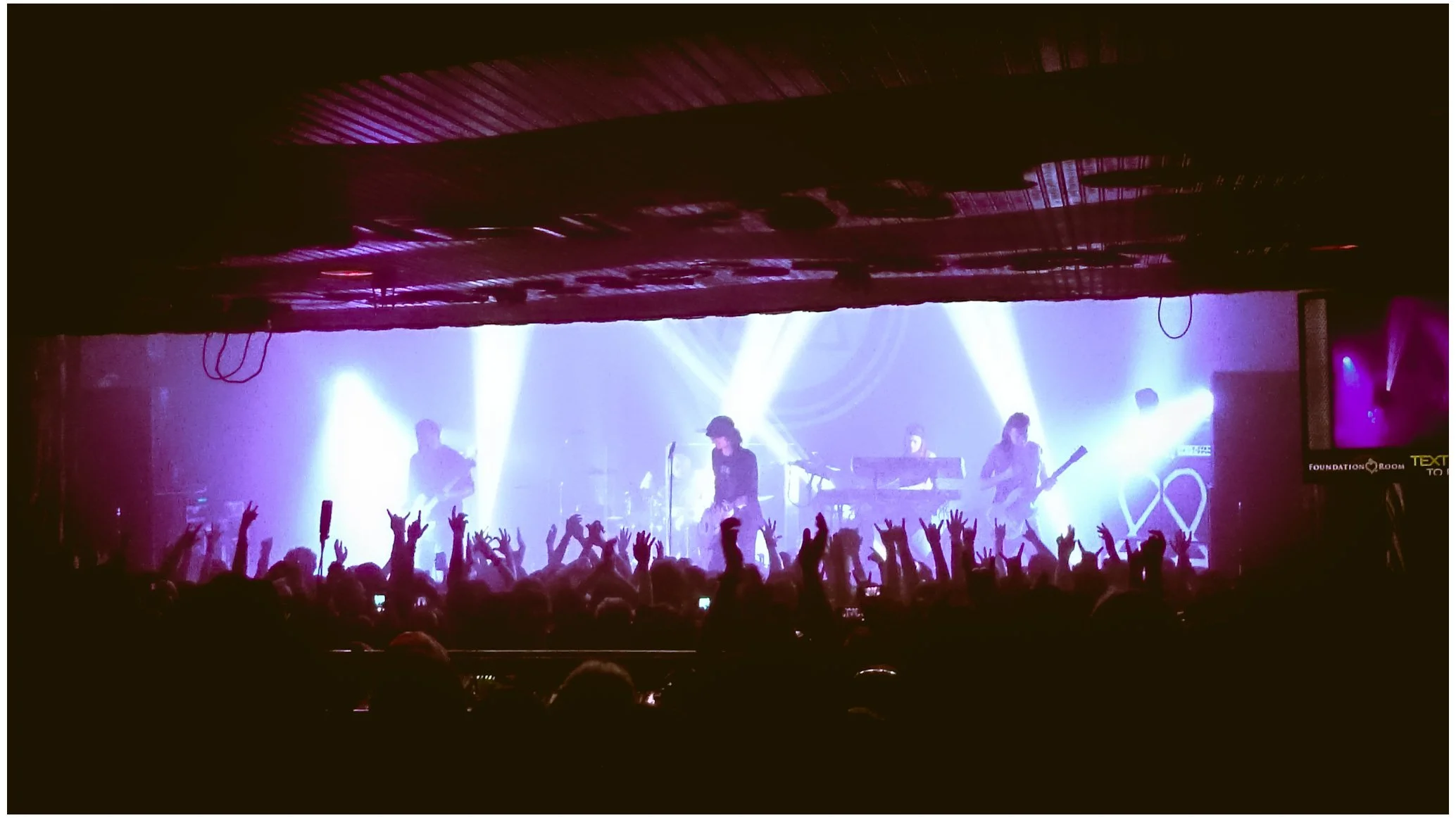 A live concert with a band performing on stage, illuminated by bright purple and white lights, with an audience raising their hands and taking photos.