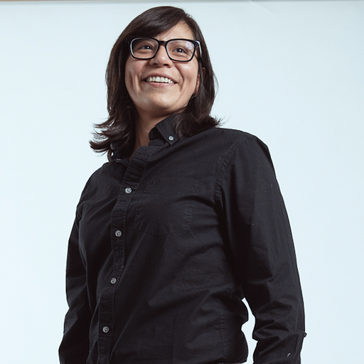 A smiling woman with dark hair, wearing glasses and a black button-up shirt, standing against a plain light background.