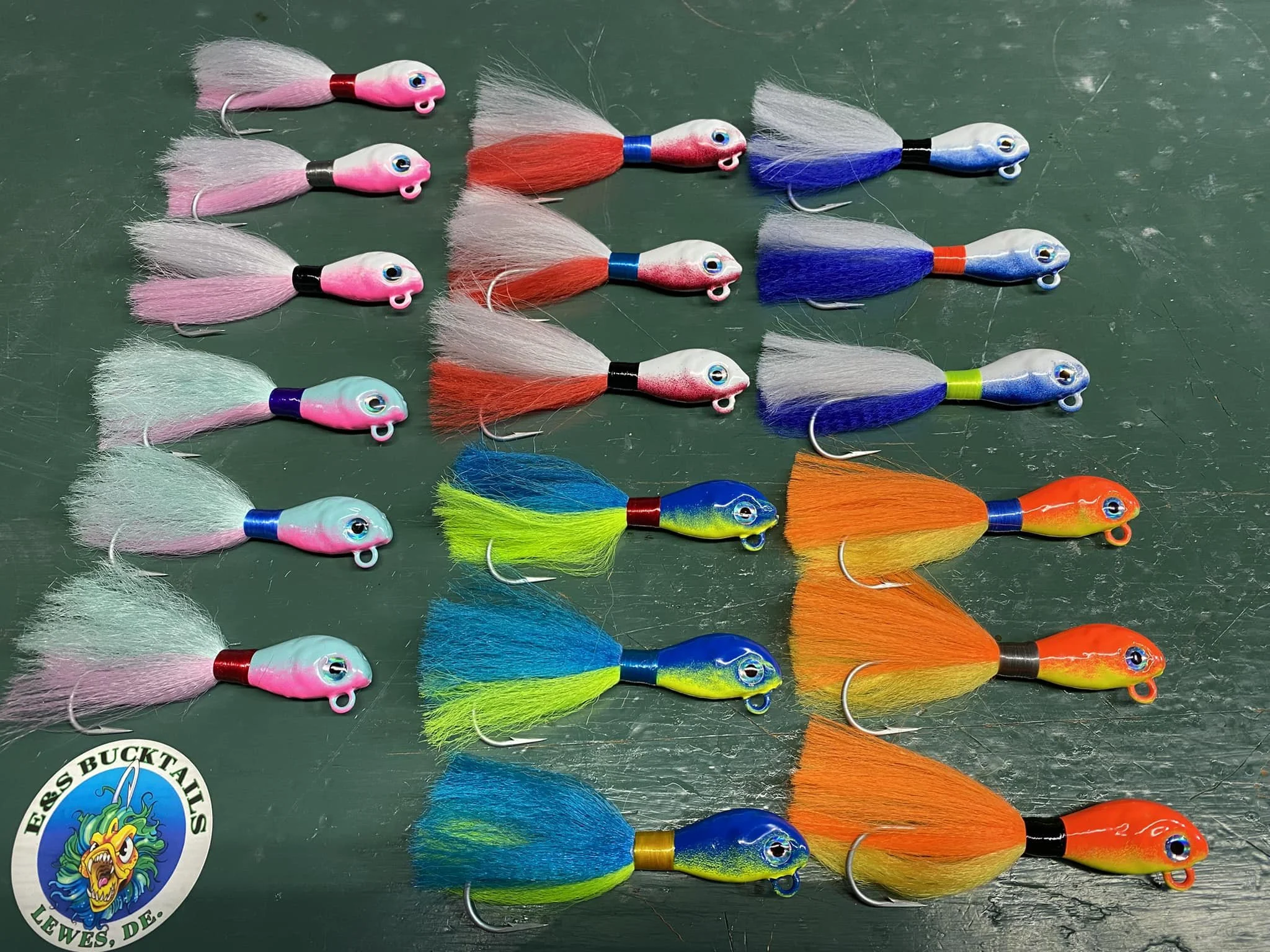 Collection of colorful fishing lures with fish-shaped heads and elongated tails arranged on a dark surface, with a logo reading 'E.S. BUCKTAILS LEWES, DE' in the bottom left corner.