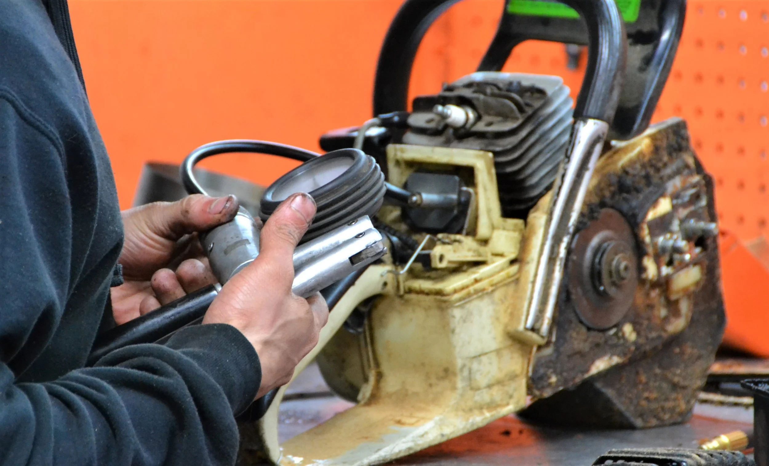 Person repairing chainsaw engine, holding a piston and carburetor, with the chainsaw's engine and rusty components visible.