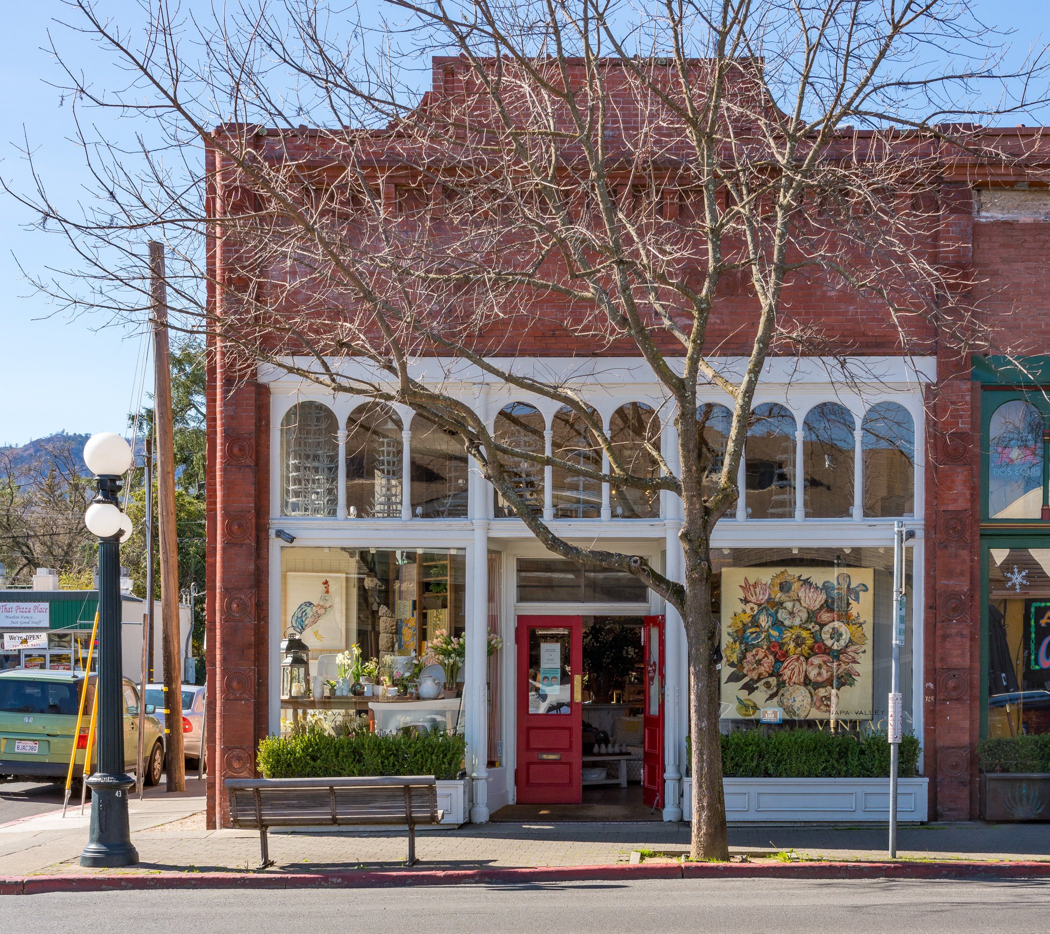 Front view of a vintage-style shop with large display windows, red door, and decorative archways, with a leafless tree in front and parked cars on the street.