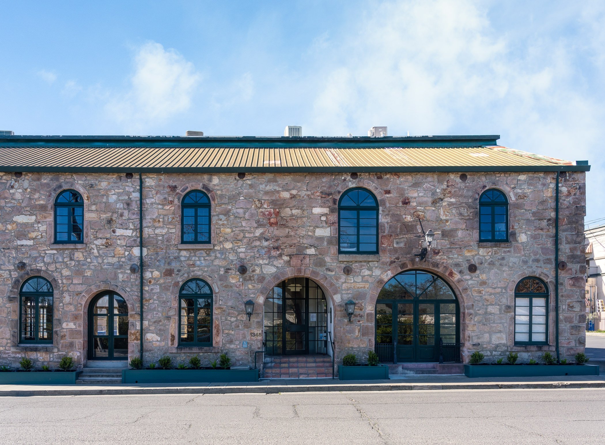 A two-story stone building with arched and rectangular blue-tinted windows, a yellow metal roof, and a glass door entrance, situated along a street with sidewalk planters and a clear sky with some clouds overhead.