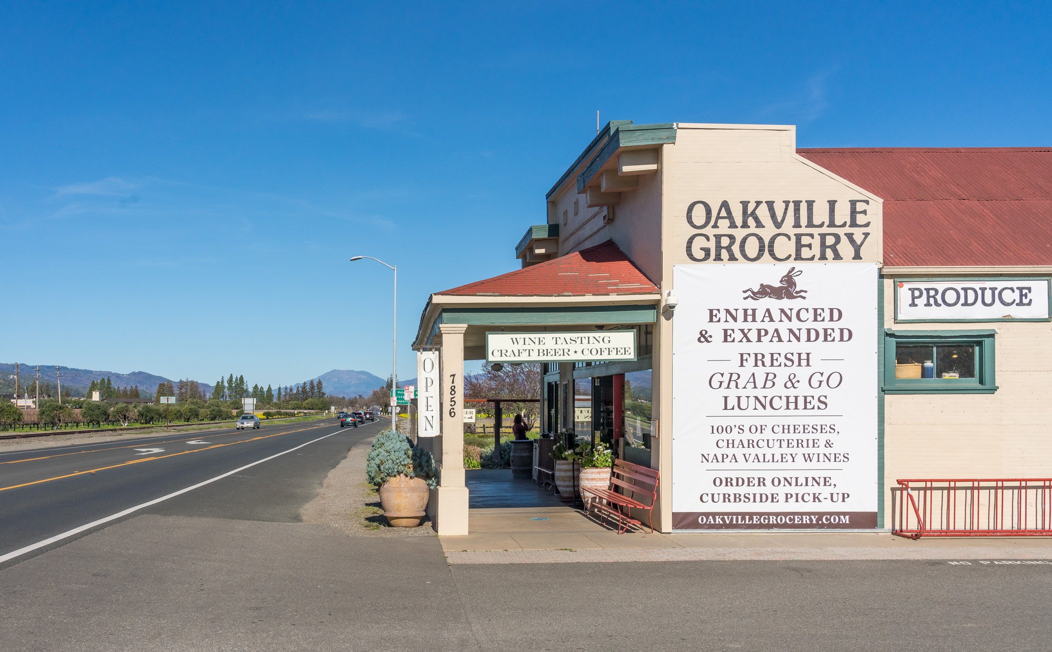 Exterior of Oakville Grocery with signs advertising wine tasting, craft beer, coffee, fresh grab-and-go lunches, cheeses, charcuterie, wines, and online curbside pickup. The building has a red roof, a porch area with a bench, and potted plants outsid