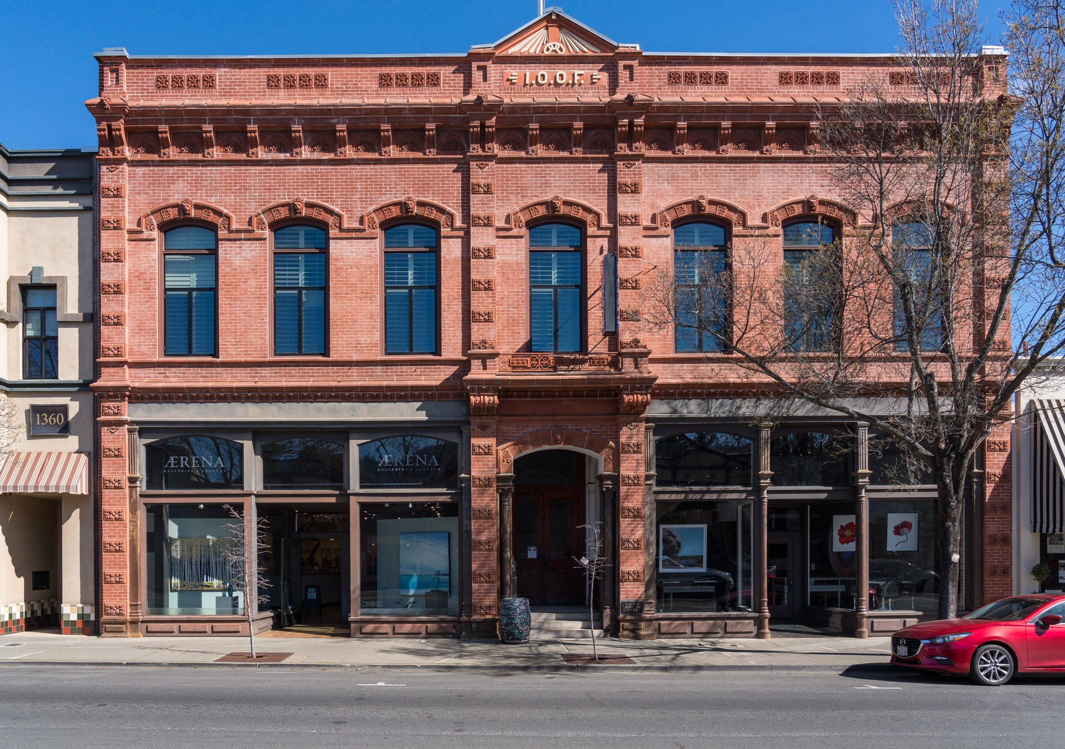 Three-story red brick building with large windows on the second and third floors, storefronts on the ground floor, a leafless tree on the sidewalk, and a red car parked in front.