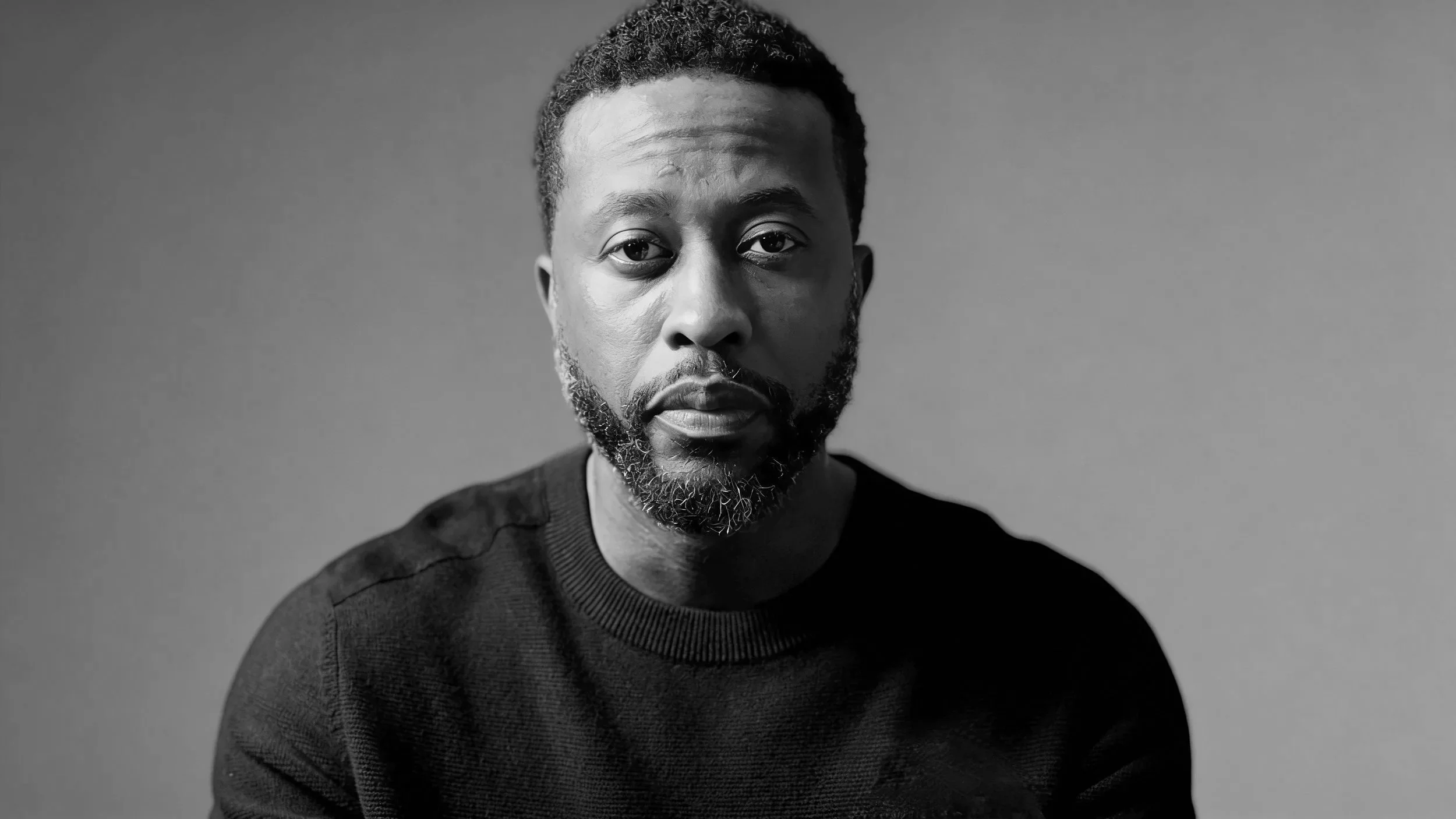 Black and white portrait of a man with a short beard and mustache, wearing a dark sweater, looking directly at the camera with a neutral expression against a plain background.