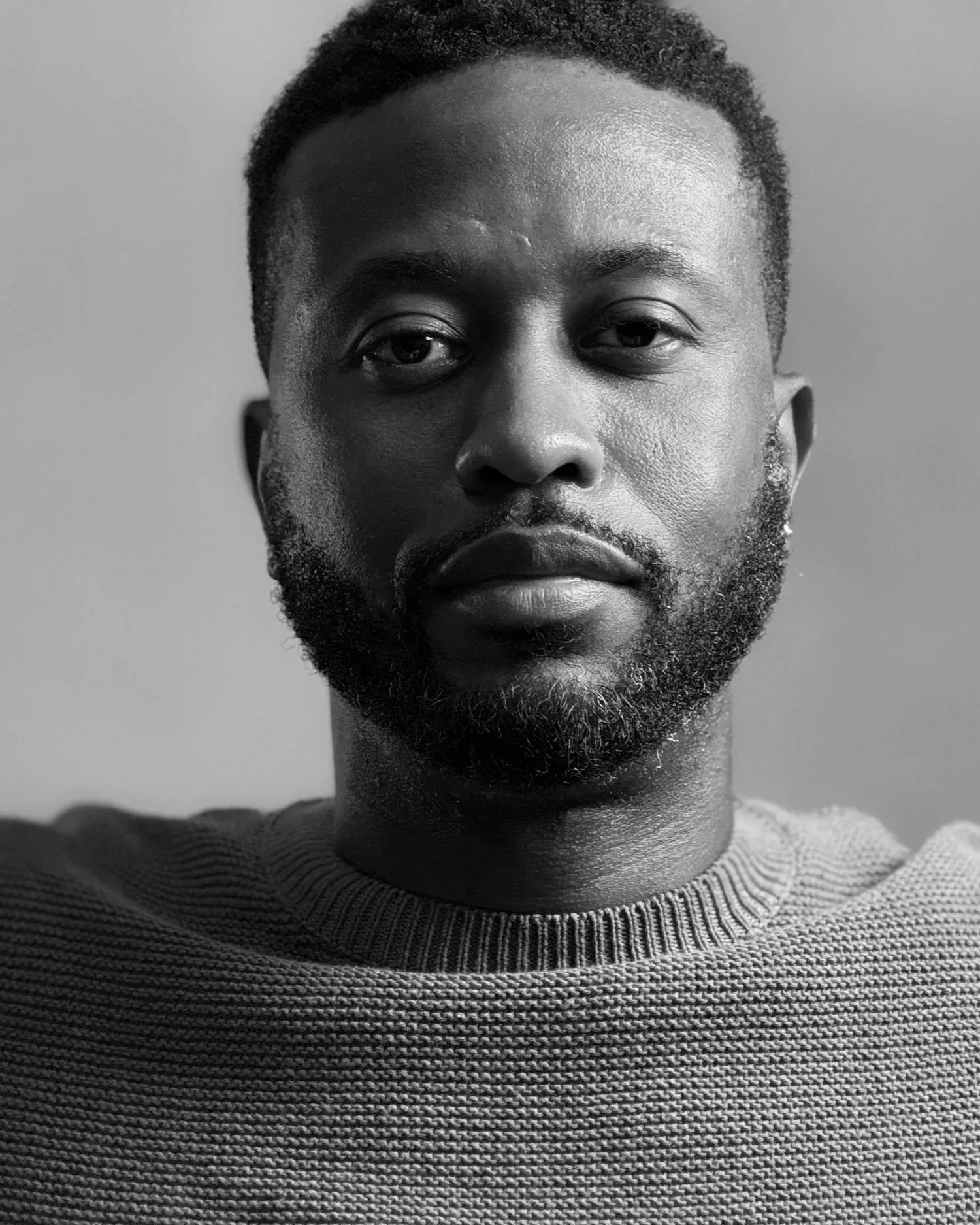 Black and white close-up portrait of a man with short, curly hair and a beard, wearing a striped sweater, looking directly at the camera.