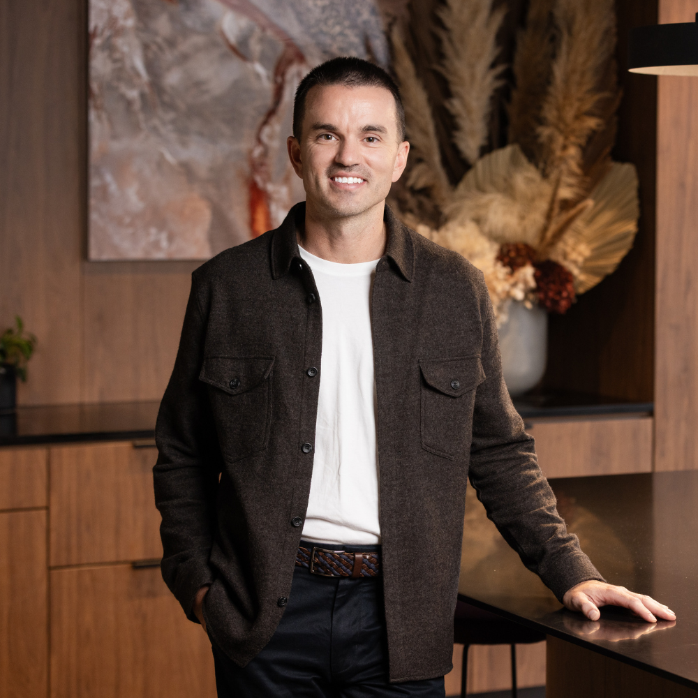 Man standing indoors near a wooden table with one hand resting on it, smiling, with a background of abstract art and dried flowers arrangement.