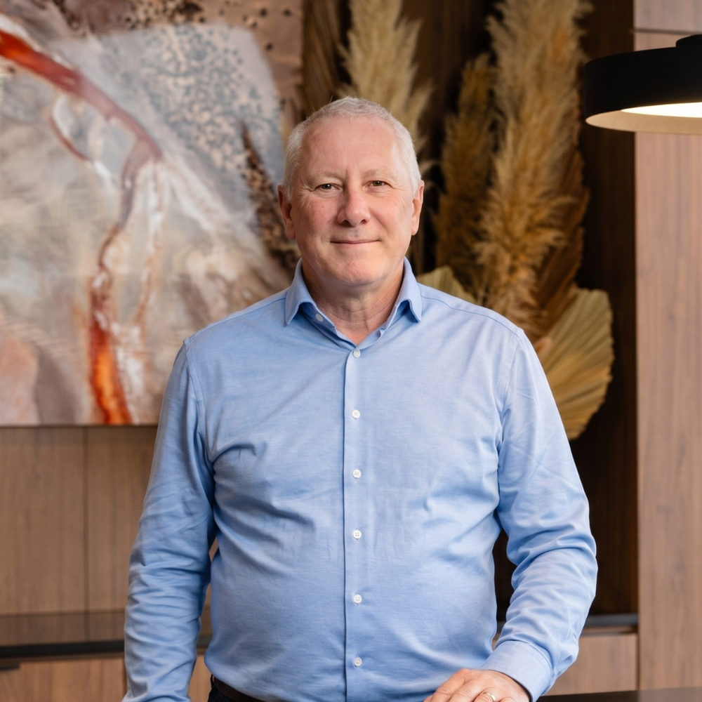 A middle-aged man with gray hair wearing a light blue dress shirt standing indoors in front of a decorated wall with dried plants and artwork.
