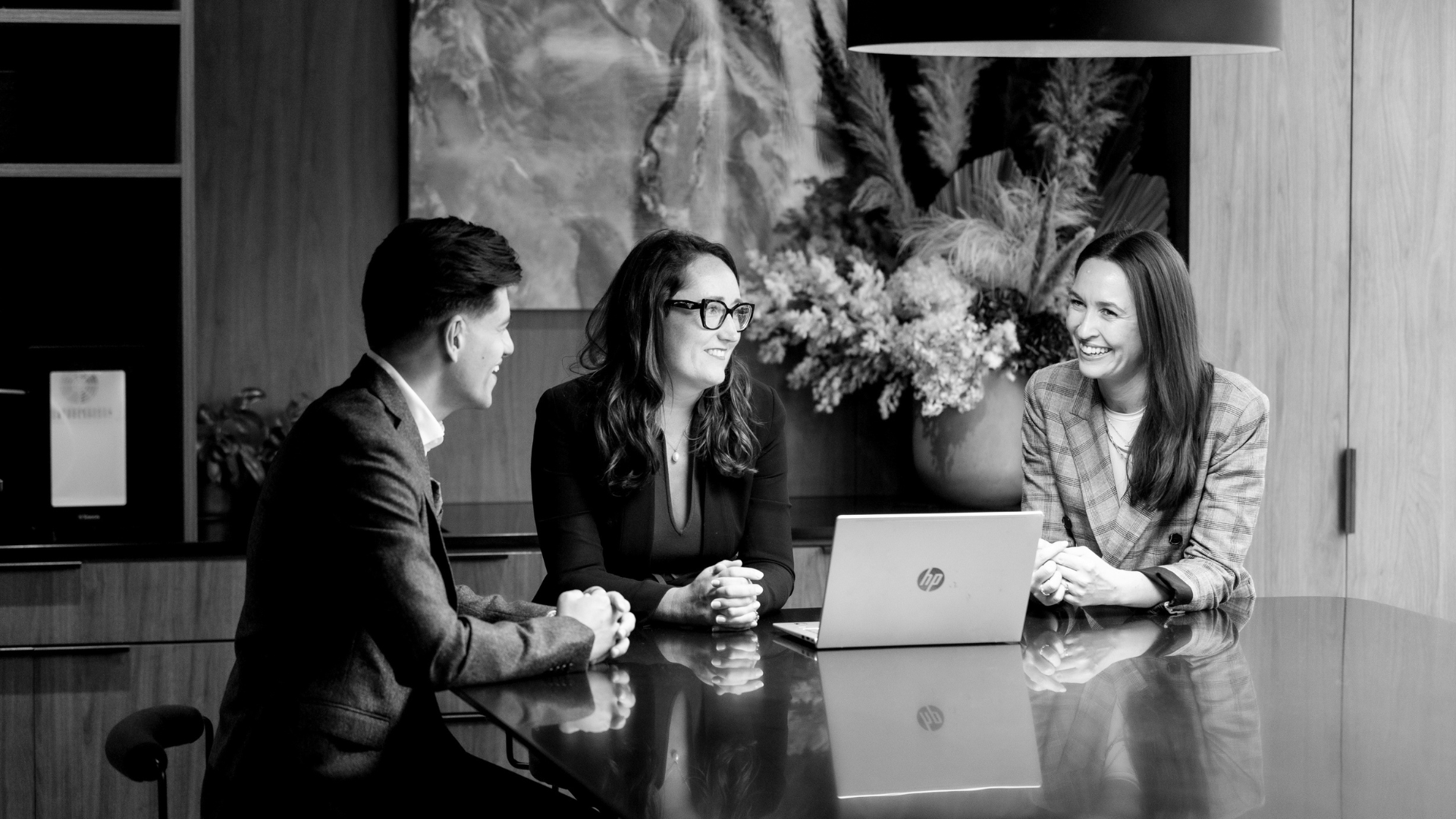 Three people are sitting at a table and having a conversation in an office setting. There is a woman with glasses and dark hair in the middle, a man with dark hair on the left, and a woman with long hair and a blazer on the right, all smiling and appearing engaged. A laptop, a large vase with flowers, and a painting are visible in the background.