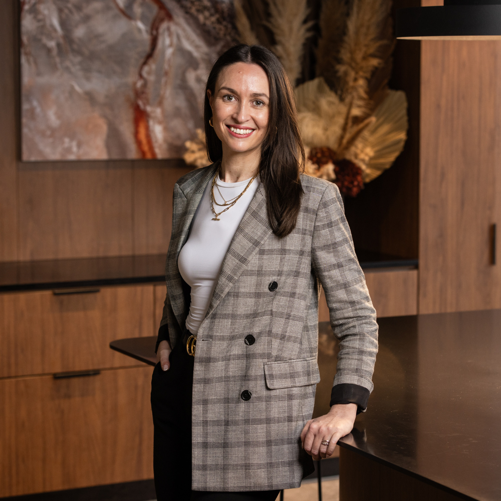 A woman in a plaid blazer smiling, standing in a modern office environment with wood paneling and artwork in the background.
