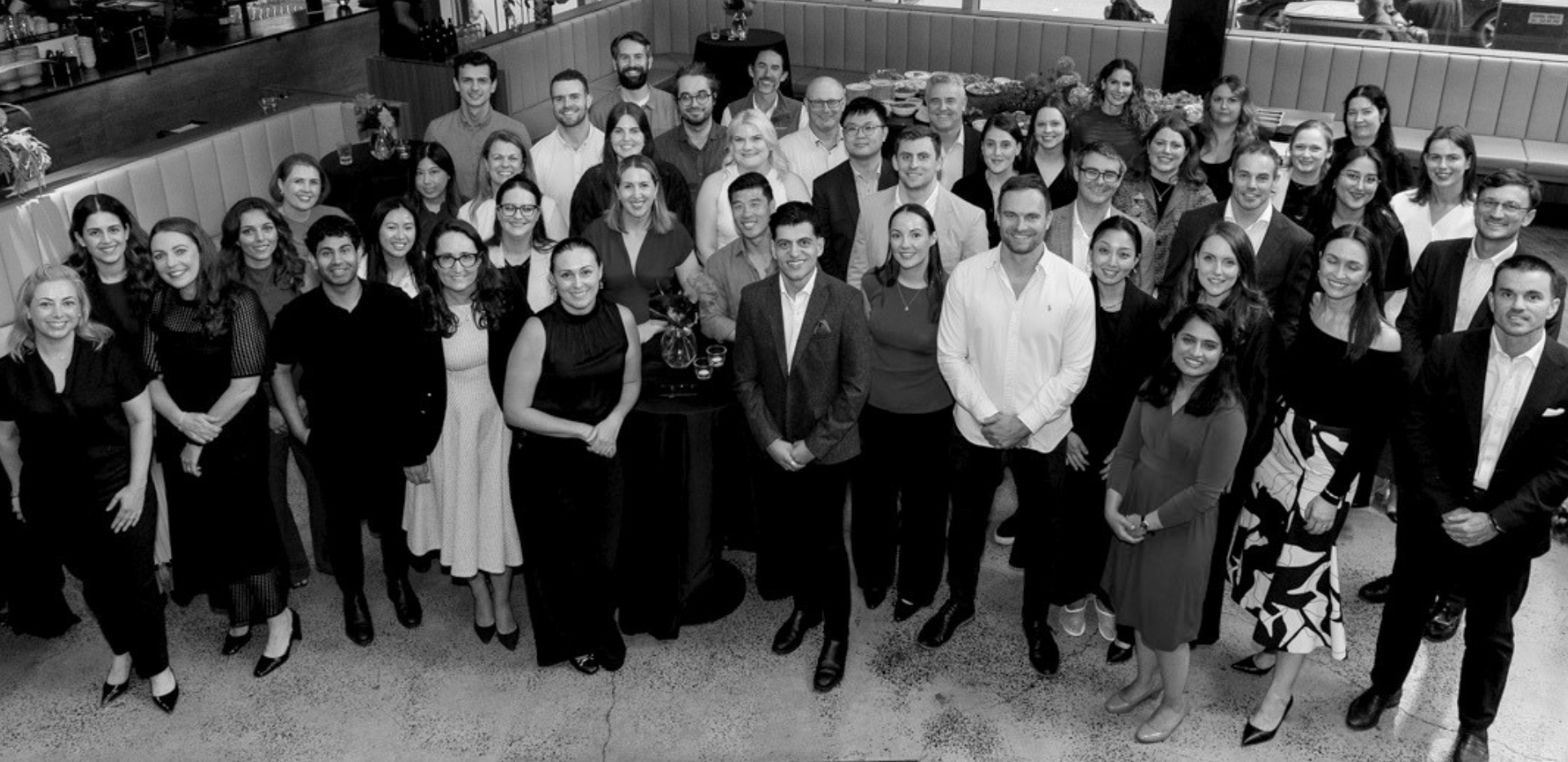 A large group of people gathered at a social event, posing for a group photo in what appears to be a restaurant or banquet hall. The group includes men and women dressed in semi-formal attire, standing and smiling for the camera with tables and food in the background.