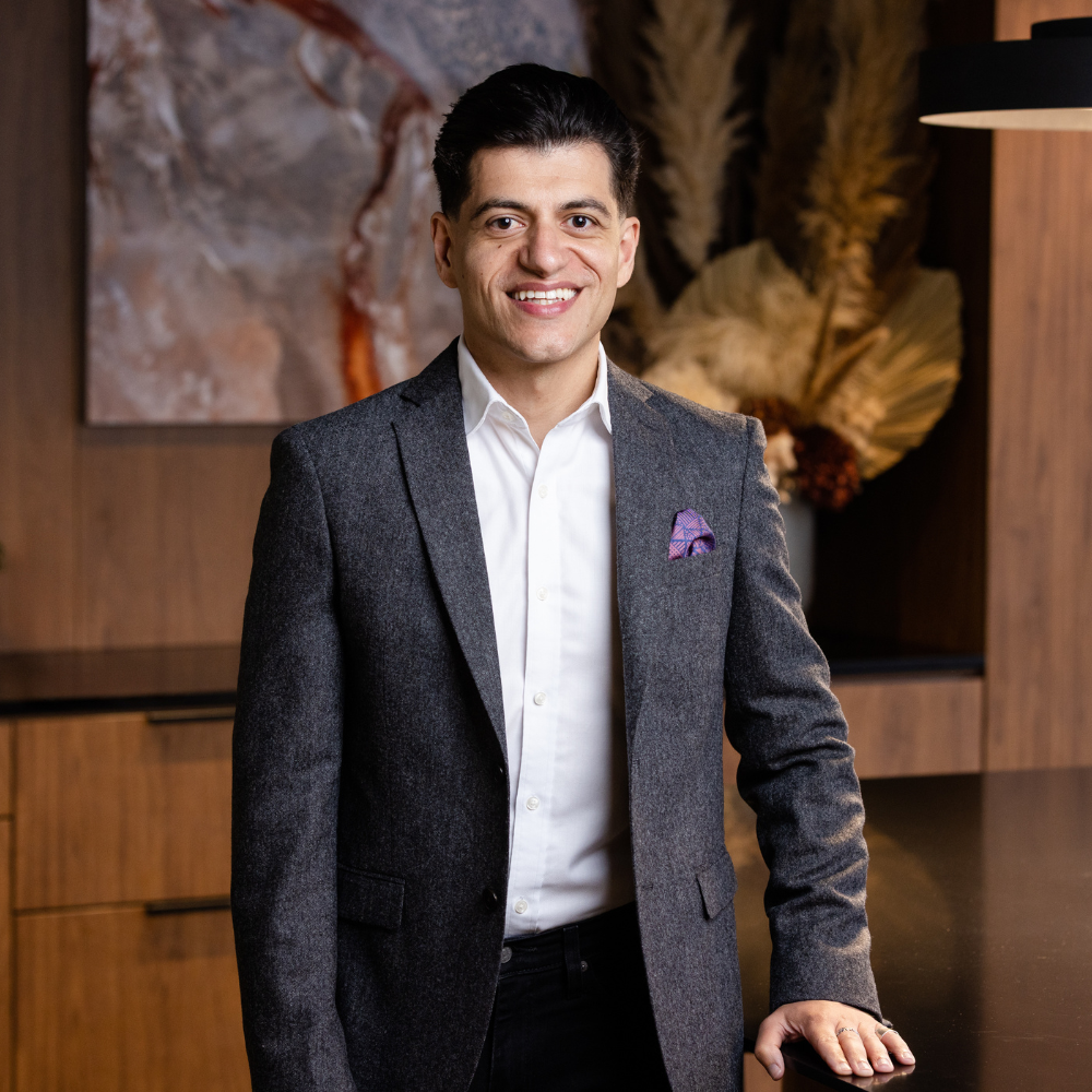 Portrait of a man in a gray blazer and white shirt smiling, standing in a room with wooden furniture and decorative dried plants.