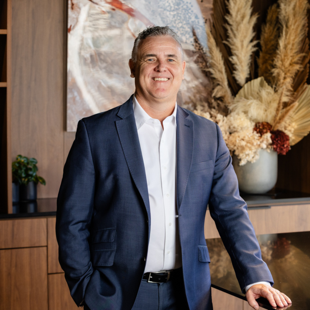 A smiling man in a navy suit and white shirt standing in a modern office with wood-paneled walls and decorative dried flowers in a large vase behind him.