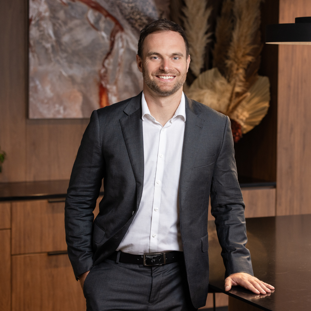 A man with a beard wearing a dark gray suit and white shirt, smiling, standing with one arm resting on a black table in a modern office or conference room.