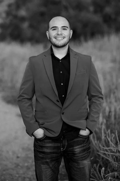 Black and white photo of a man in a blazer and jeans standing outdoors with hands in pockets and smiling.