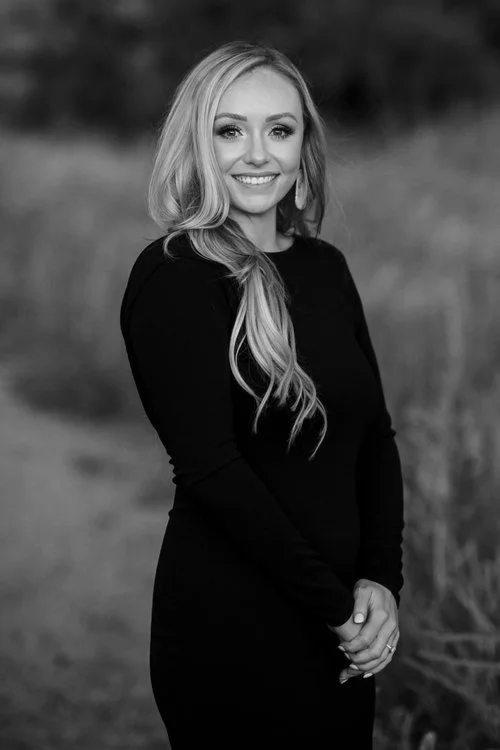 A smiling young woman with long, wavy hair wearing a black dress outdoors in a natural setting.