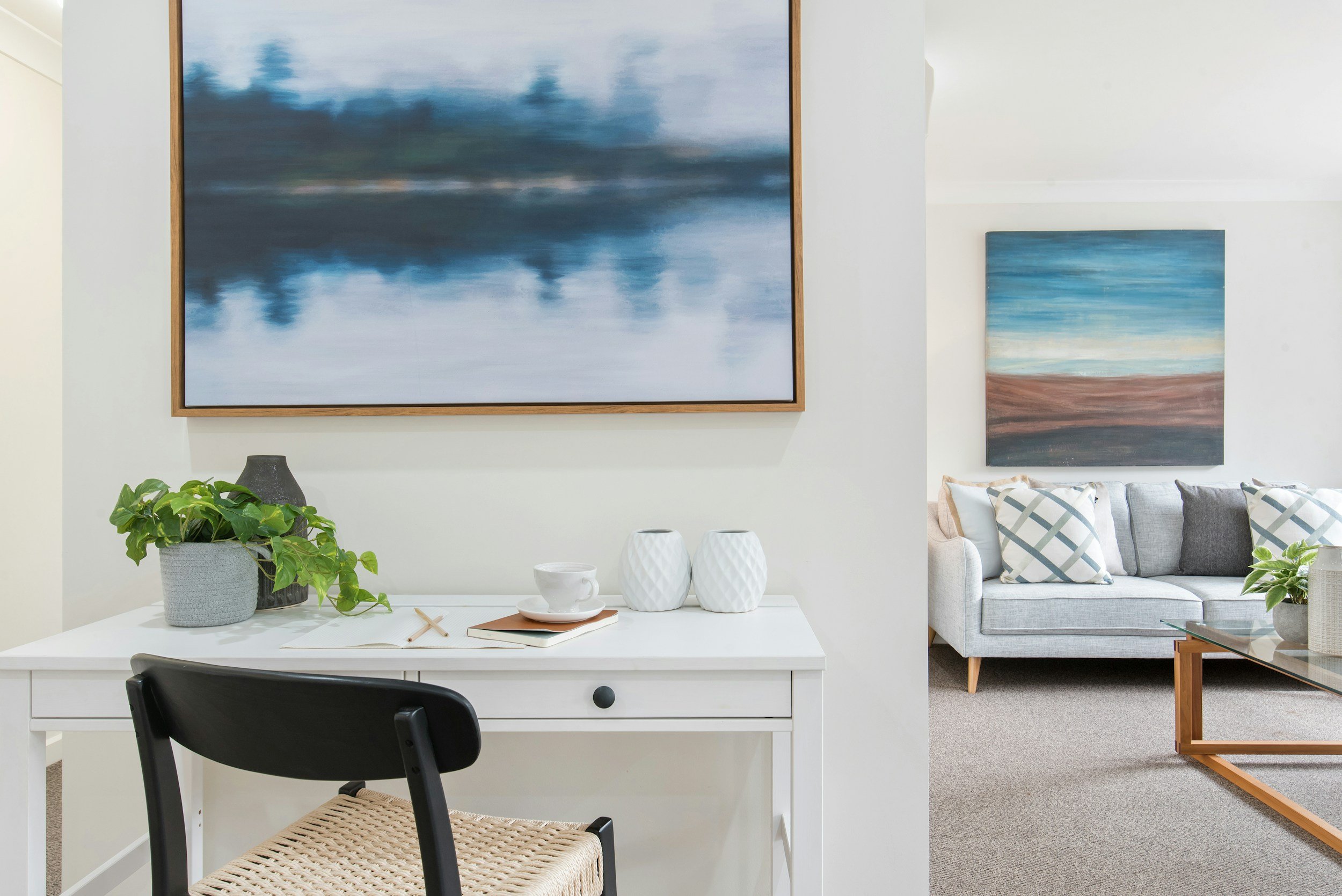 Modern living room with white desk, black chair, green plant, abstract paintings, grey sofa, and decorative pillows.