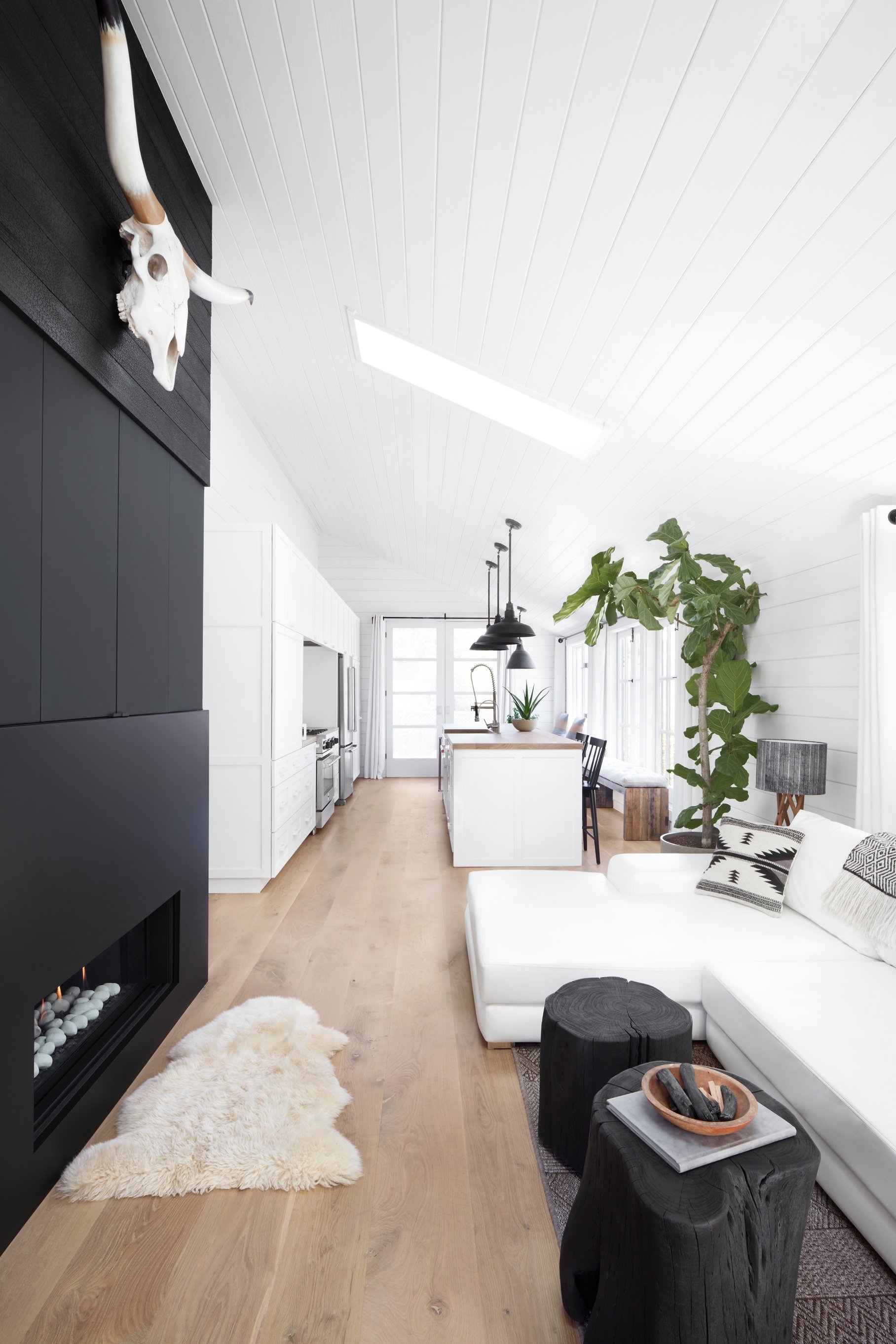 Modern living room with white walls, wood floor, black fireplace, animal skull, white sectional sofa, black stools, large plant, and open-plan kitchen.