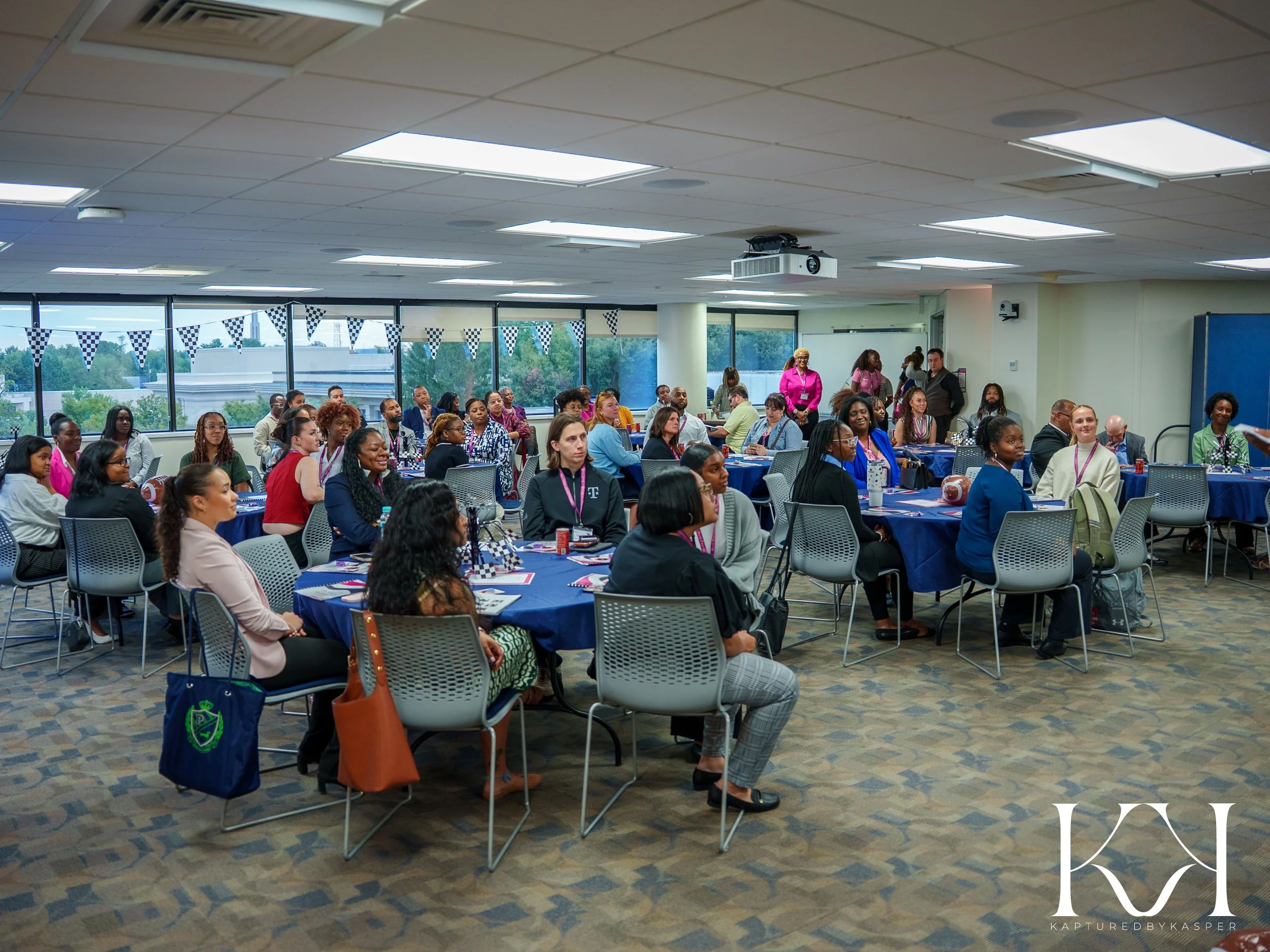 A large conference room filled with mostly women seated at round tables covered with blue tablecloths, attending a presentation or seminar. There are a few men present and some standing at the back. The room has large windows with a view of trees and a building outside, and was decorated with black-and-white checkered flags across the windows.