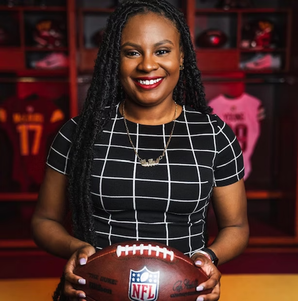 Woman in black and white checkered shirt holding NFL football, standing in front of a sports locker with football jerseys and equipment.