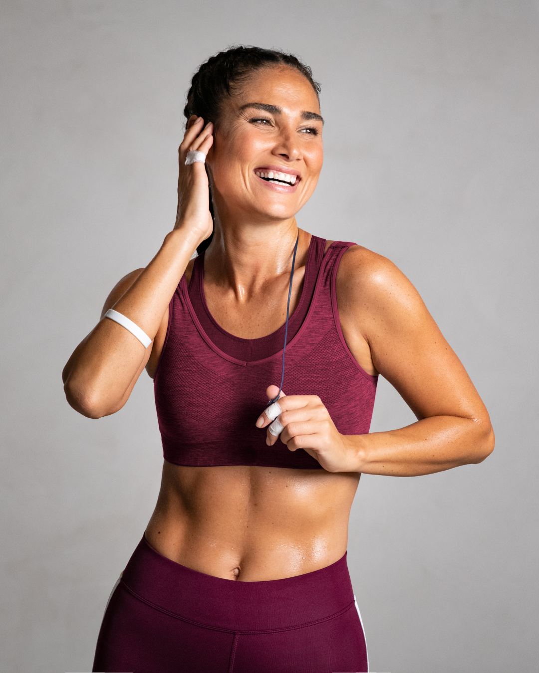 A woman in athletic wear, smiling, adjusting her earphone, with sweat on her skin.