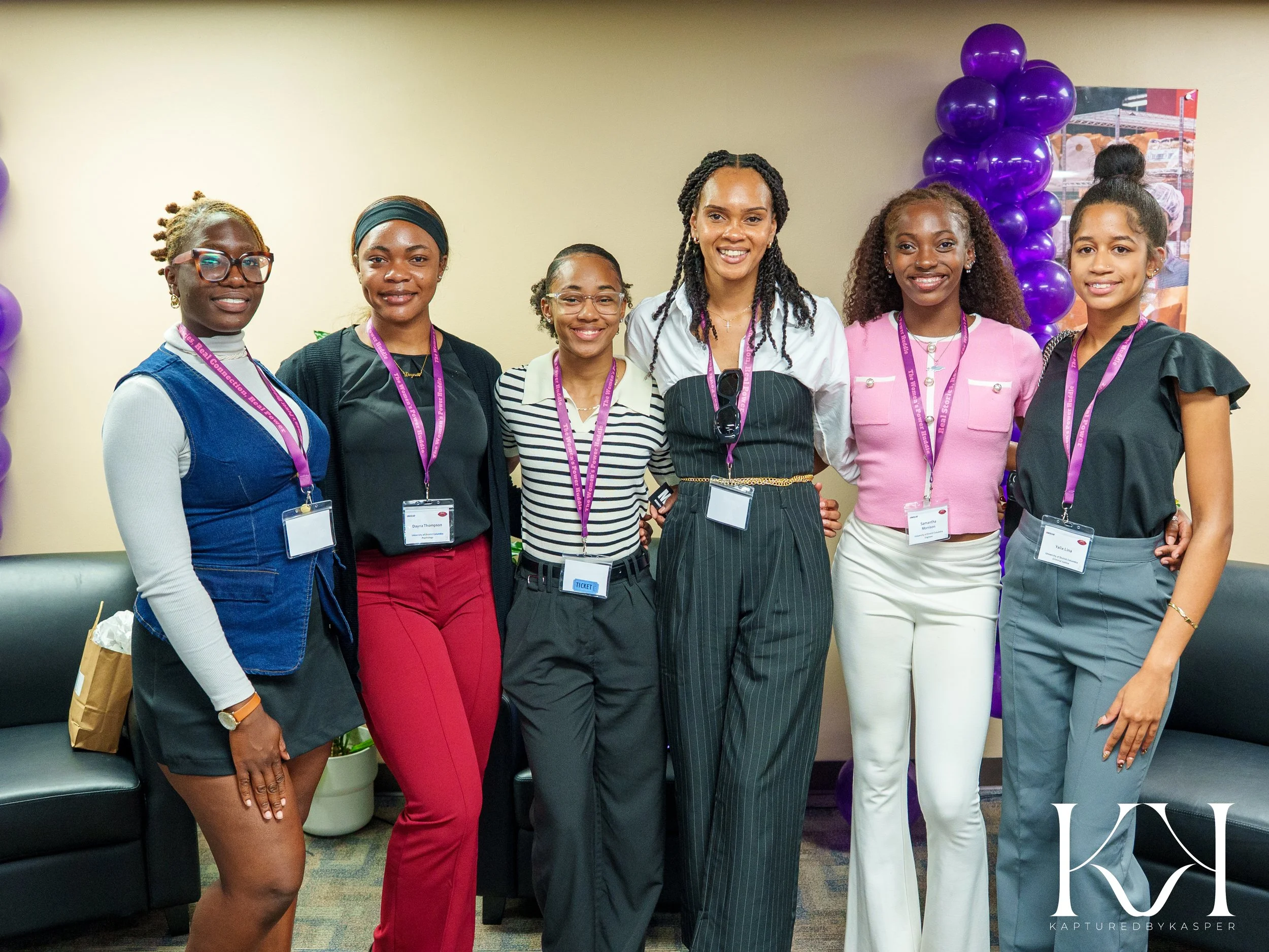 Six women smiling and standing together at an indoor event, wearing business casual clothing, with purple balloons and a poster in the background.