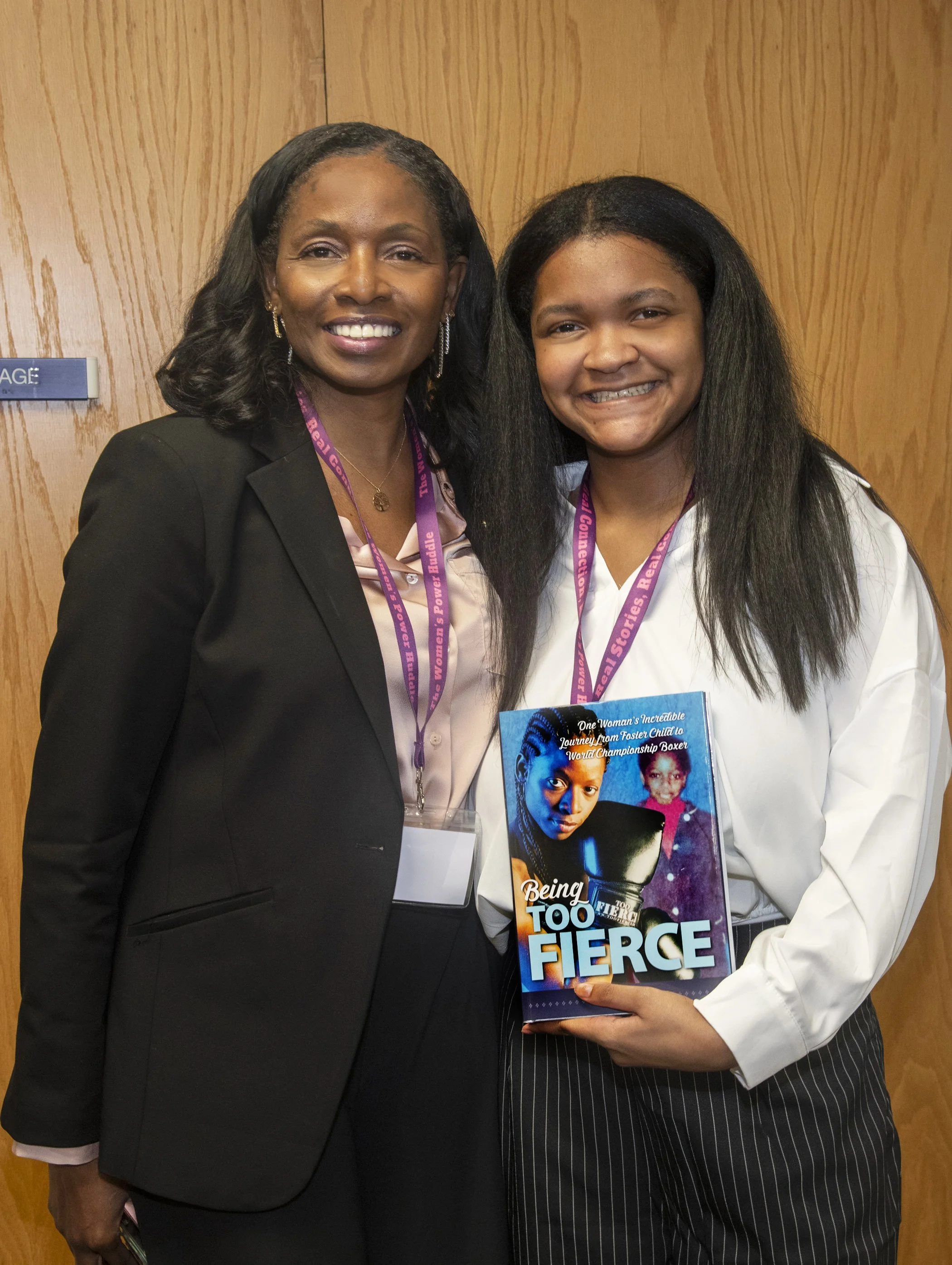 Two women standing together, smiling, with one holding a book titled 'Being Too Fierce'. They are wearing conference lanyards and standing in front of a wooden background.