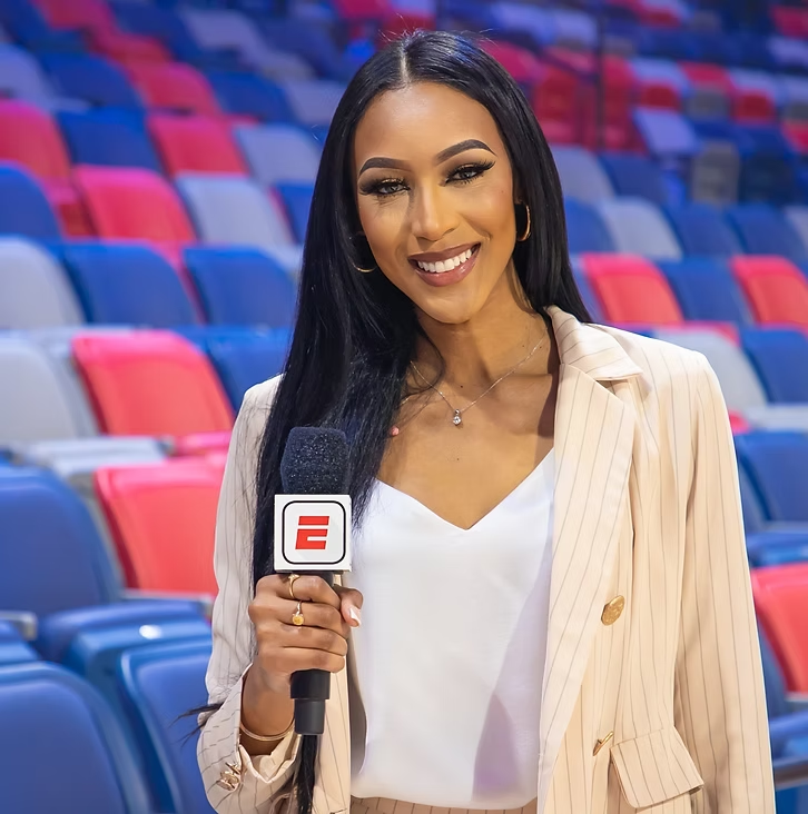 A woman with long black hair and a white shirt holding a microphone with ESPN logo, standing in front of rows of red, blue, and gray stadium seats.