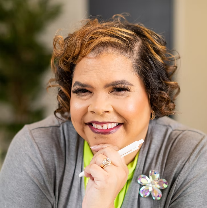 A smiling woman with curly hair holding a pen near her chin, wearing a gray top and a decorative flower pin.