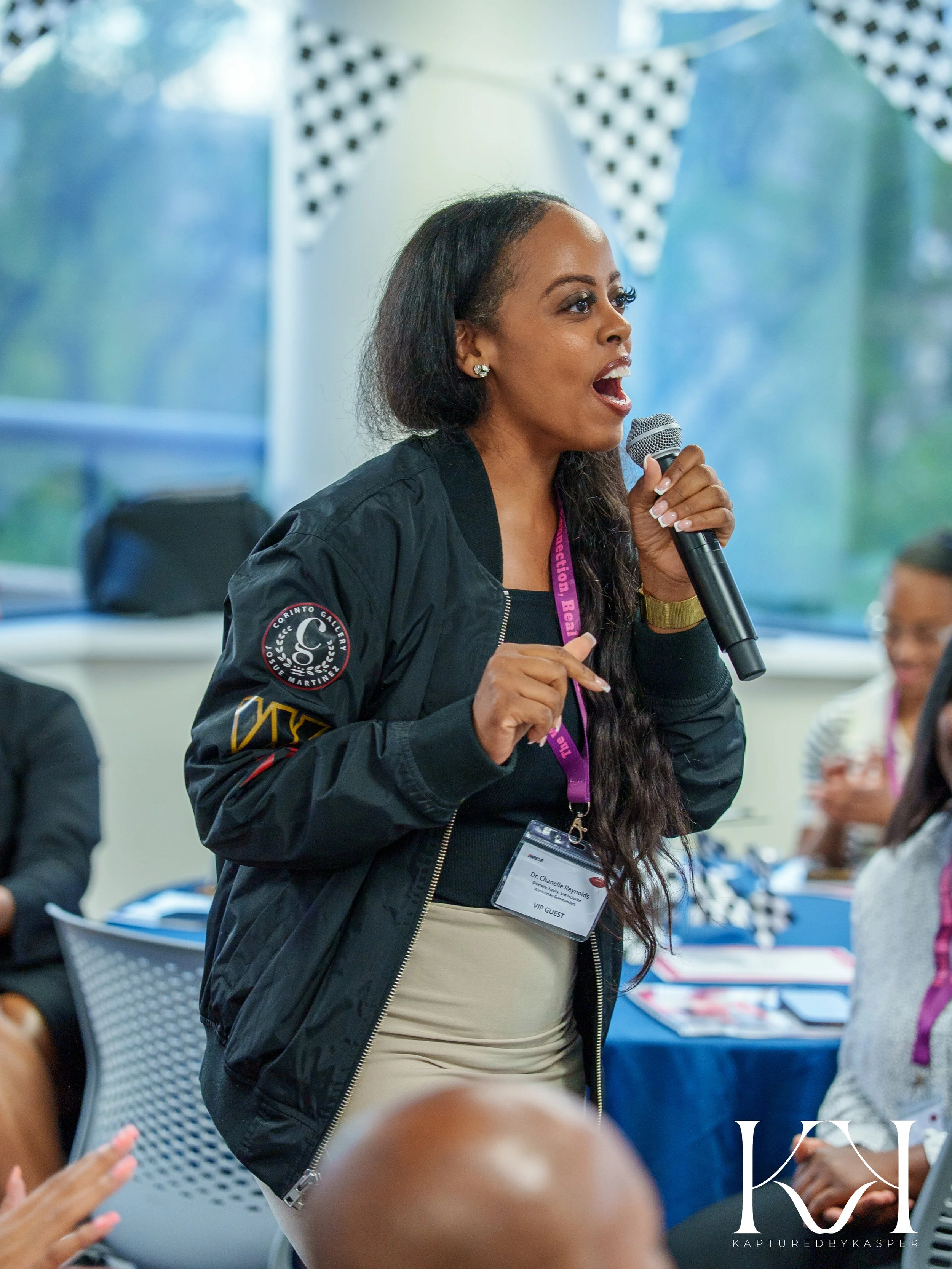 A woman speaking into a microphone at a conference, wearing a black bomber jacket with patches and a VIP guest badge. She is standing in front of an audience, with other attendees reseated at tables in the background.