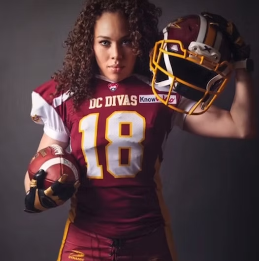 A female football player wearing a maroon and gold uniform with the number 18 and the words 'DC Divas' on the front, holding a football in her left hand and a helmet in her right hand.